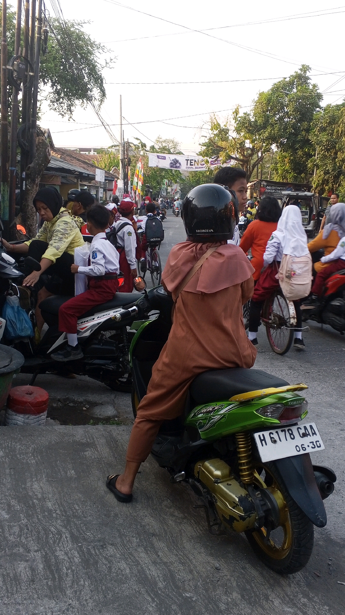In Semarang, Indonesia, on a paved street lined with buildings and trees, a group of people are gathered, many on motorbikes. In the foreground, a person wearing a brown hooded robe and a black helmet sits on a green and gold Yamaha scooter. Their feet are clad in black sandals. The scooter's license plate reads "H 6178 CAA 06-30". Behind them, several other individuals are visible, including some wearing school uniforms. A young boy in a white shirt and red pants, also on a scooter, holds a stack of white papers. A banner with the word "TENGA" and an image of a blueberry is displayed in the background. The scene appears to be during daylight.