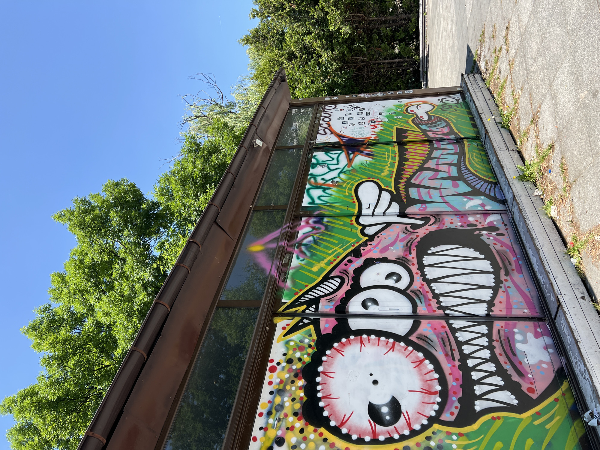 The image shows a building with graffiti on its walls. The graffiti is colorful and features cartoonish characters. The building appears to be located outdoors, possibly in a park or public space, with trees and a blue sky in the background. The pavement in front of the building is made of concrete slabs. The building itself has a brown roof and dark windows.