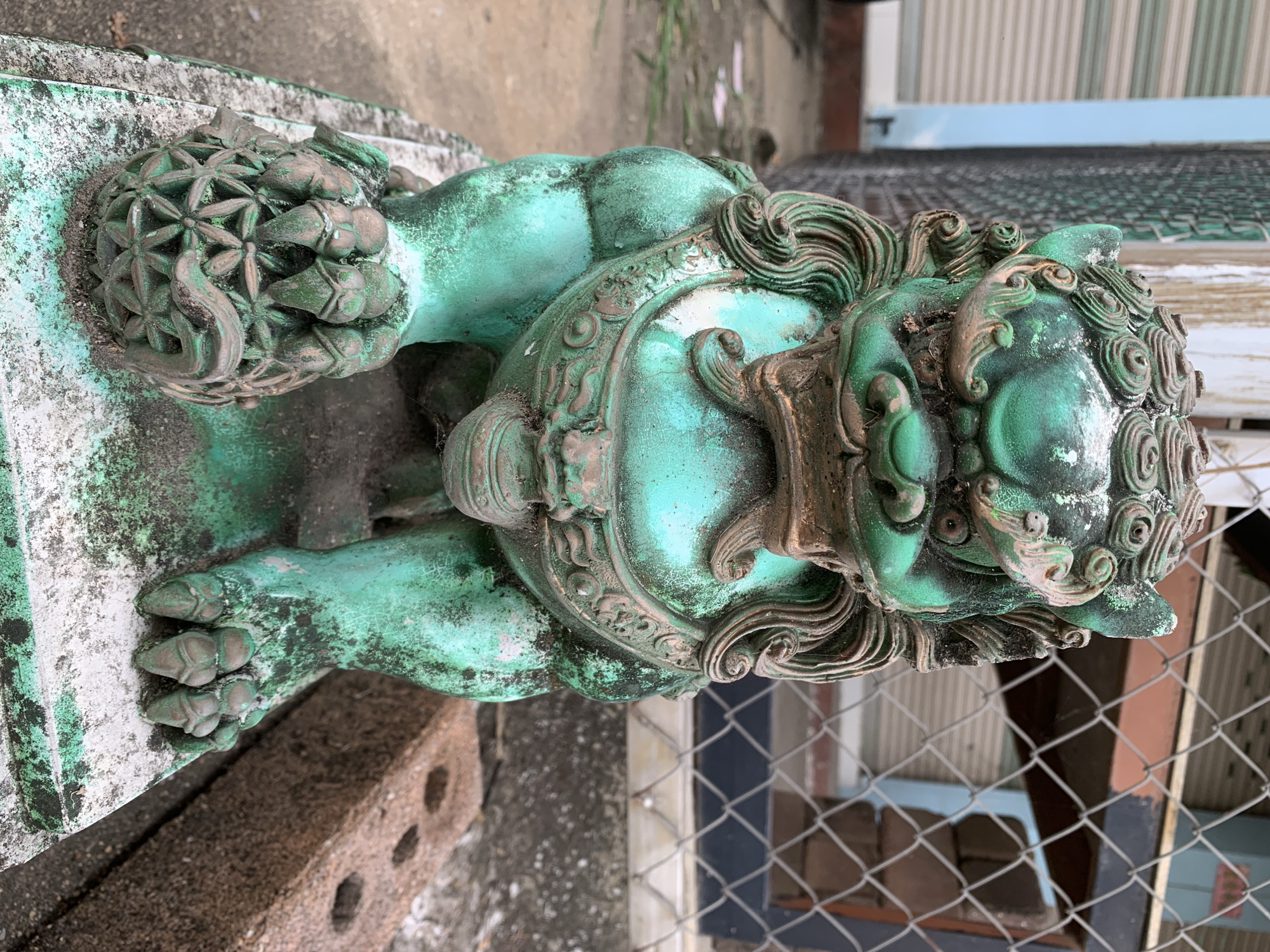 A weathered, green-patinated statue of a Foo dog, likely a guardian lion, stands on a concrete platform outdoors. The statue is detailed, with a mane, ornate collar, and one paw resting on a decorative sphere. The green patina is chipped and weathered, revealing underlying bronze or copper tones in places. The statue is situated in what appears to be a back alley or outdoor storage area. In the background, a chain-link fence, a corrugated metal structure, and a brick are visible. The ground is paved with concrete, and some debris is scattered around. The lighting suggests it is daytime. The location is identified as Ban San Ton Kok, Thailand. No people are present in the image.