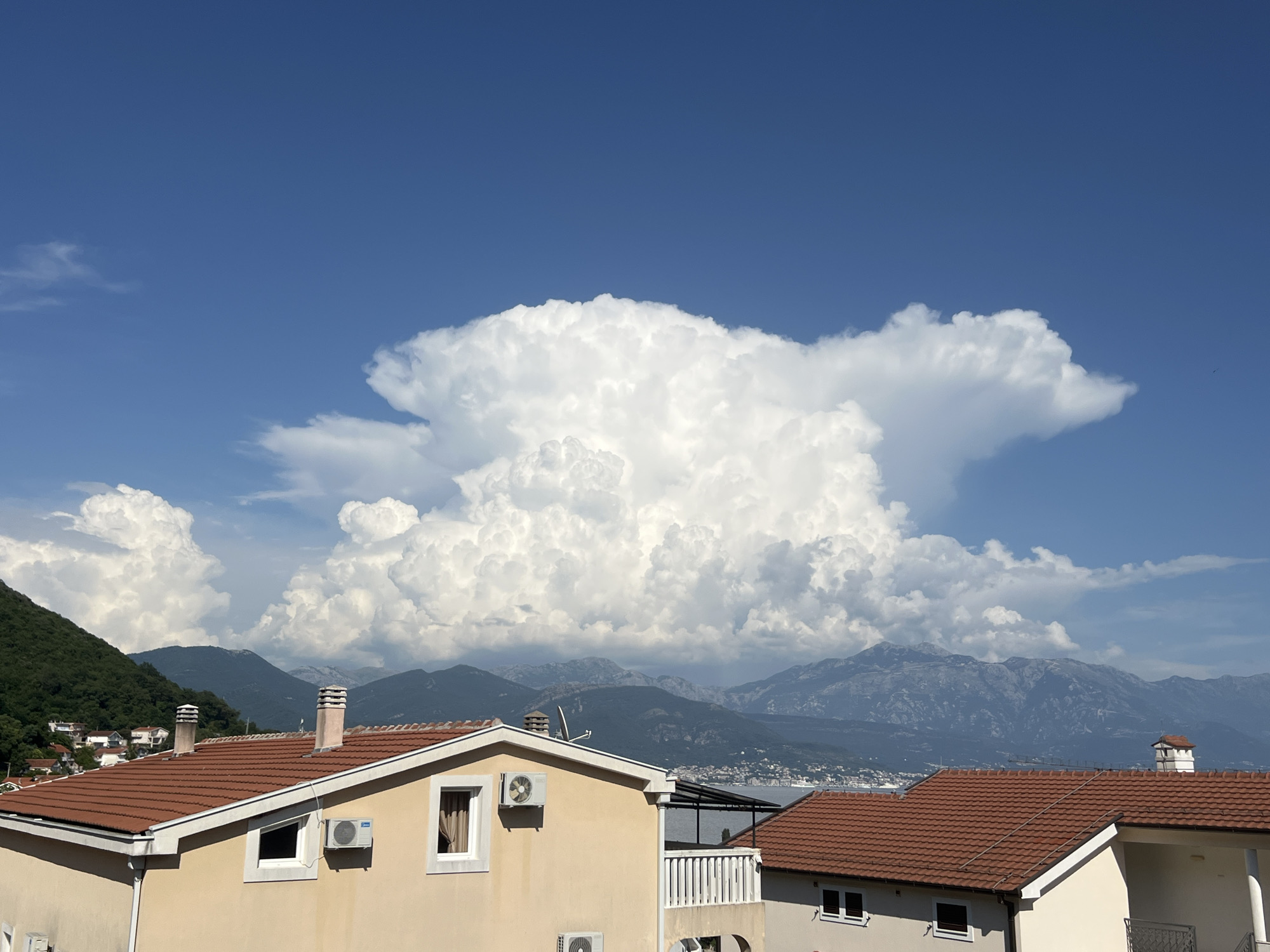 This is a medium-shot, outdoor, daytime photograph taken from a high vantage point, looking out over a town nestled between a body of water and a mountain range under a bright blue sky with dramatic, large cumulus clouds. The foreground is dominated by the tiled roofs of two houses. The house on the left has a pale yellow facade, a red-tiled roof, and features two windows, an air conditioning unit, and a chimney with a striped cap. The house on the right also has a red-tiled roof and shows a portion of its facade with windows. In the middle ground, a densely vegetated hillside with scattered houses leads down to the water. The background features a broad expanse of water, beyond which a vast, rugged mountain range stretches across the horizon. The sky is a deep blue, punctuated by large, towering white clouds, particularly a massive formation in the center of the frame that resembles a cauliflower or a mushroom cloud, suggesting potential stormy weather further inland. The scene is one of natural beauty with man-made structures integrated into the landscape, likely depicting a residential area in Baošići, Montenegro, given the characteristic combination of coastal, mountainous, and Mediterranean architectural elements. There are no people visible in the frame.