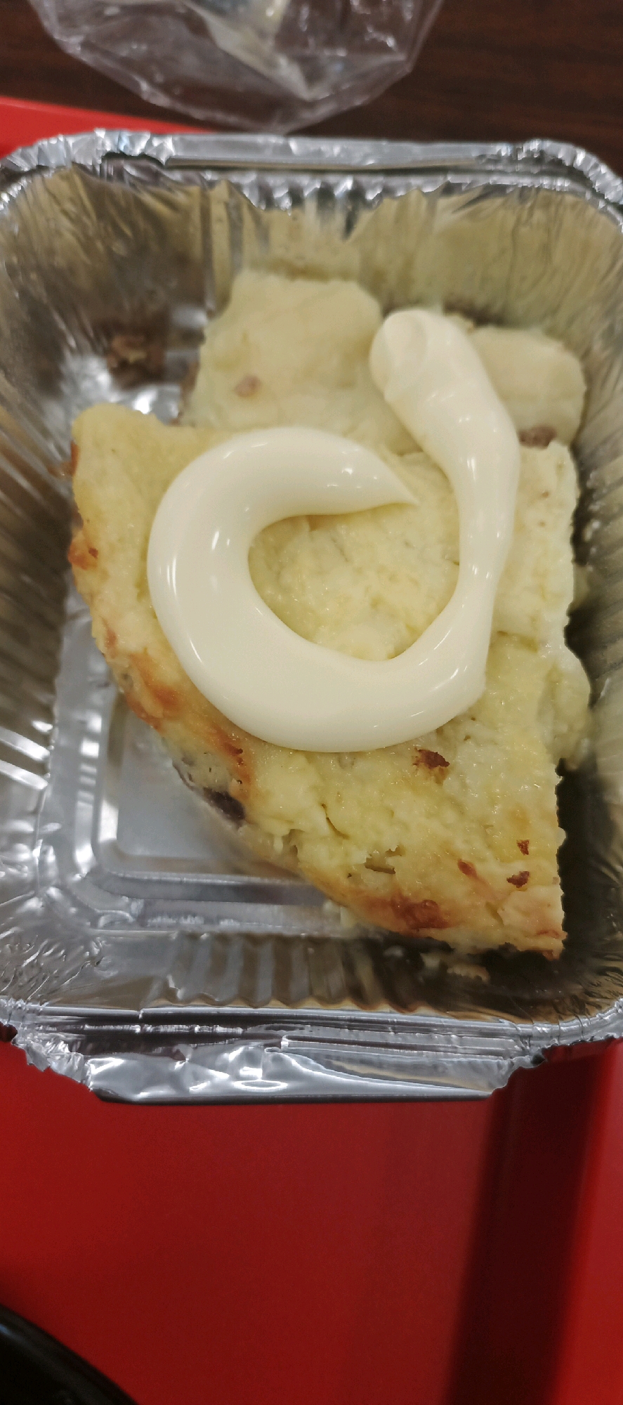 A close-up, overhead shot shows a slice of what appears to be a savory pie or casserole, likely made with mashed potatoes, in a rectangular aluminum foil container. The dish has a golden-brown crust and is topped with a swirl of a creamy white sauce, possibly mayonnaise or a similar dressing. The container is positioned on a red surface, and a portion of a crumpled plastic wrapper is visible in the upper background. The scene is static, with no people or visible actions. The lighting is diffused, casting soft shadows. The image is consistent with a prepared food item meant for consumption.