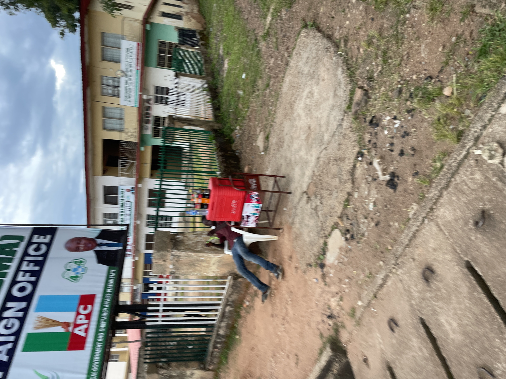 The image captures a street scene in Jos, Nigeria, likely in front of a political campaign office, evidenced by a large banner visible in the background with "CAMPAIGN OFFICE" and the APC logo. A man, dressed in a maroon shirt and blue jeans, is seen lounging in a white plastic chair, appearing to be relaxing or taking a break. In front of him is a red cooler filled with various bottled drinks, including Coca-Cola, suggesting he is a vendor. The surrounding environment appears to be a mix of paved and unpaved ground with some sparse vegetation and scattered debris. The buildings in the background suggest an urban or semi-urban setting. The overall impression is one of a casual street-side business operation in a somewhat unkempt urban area.