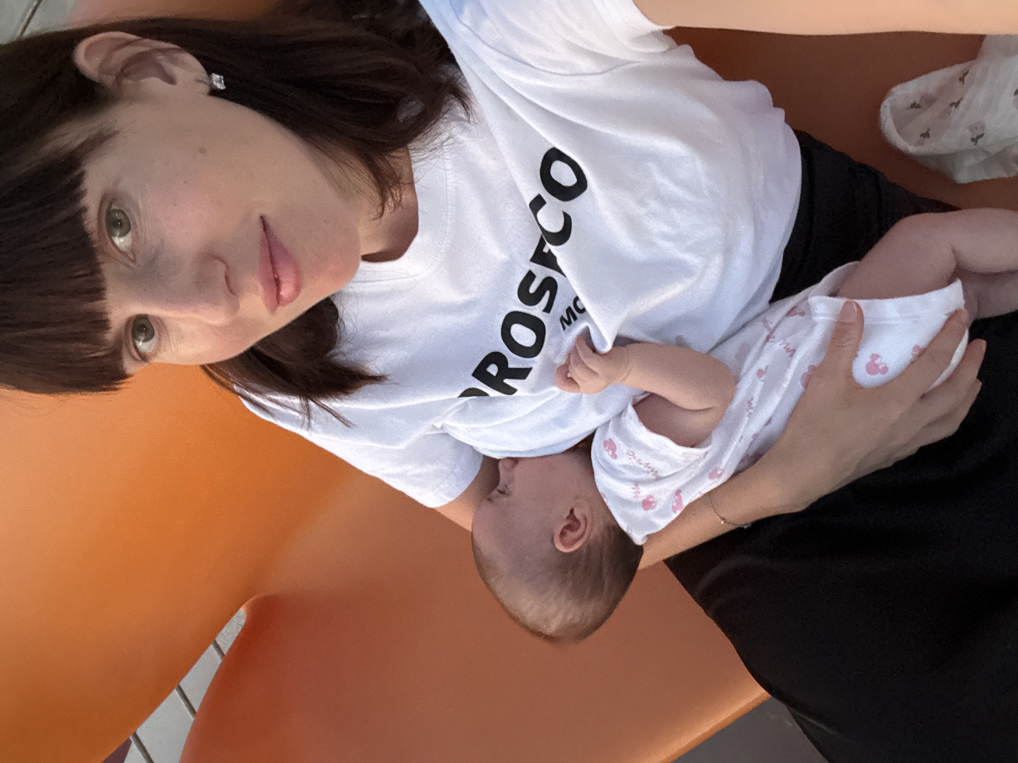 A medium shot photo captures a woman breastfeeding a baby. The woman has dark hair with bangs and is wearing a white t-shirt with black text that reads "ROSFCCO". She is wearing a silver stud earring. The baby is wearing a white onesie with a pink floral pattern. The background appears to be an outdoor setting with an orange structure and a hint of a railing. The photo was taken in the city of Isola delle Femmine, Italy.