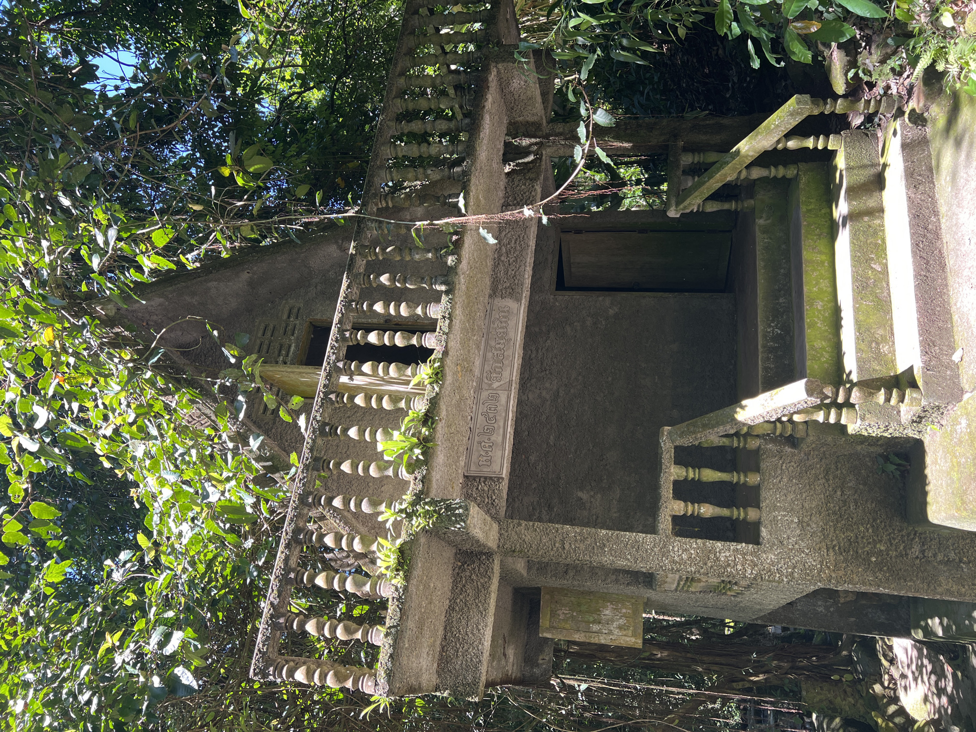 A concrete structure, possibly a shrine or small temple, is nestled within a lush, tropical environment. The building is constructed of weathered concrete, with a small balcony featuring a balustrade made of decorative concrete posts. Greenery, including vines and small plants, grows on and around the structure, indicating it is integrated into the natural landscape. Steps lead up to the building, and a wooden door is visible. Above the door, a plaque with Thai script is affixed to the facade. The scene is characterized by the interplay of man-made architecture and dense vegetation, suggesting a location in a natural setting, likely a temple complex or a secluded area. The presence of abundant foliage, dappled sunlight filtering through leaves, and the weathered appearance of the concrete all contribute to a sense of age and integration with nature. This structure is located in Baan Thurian, Thailand.