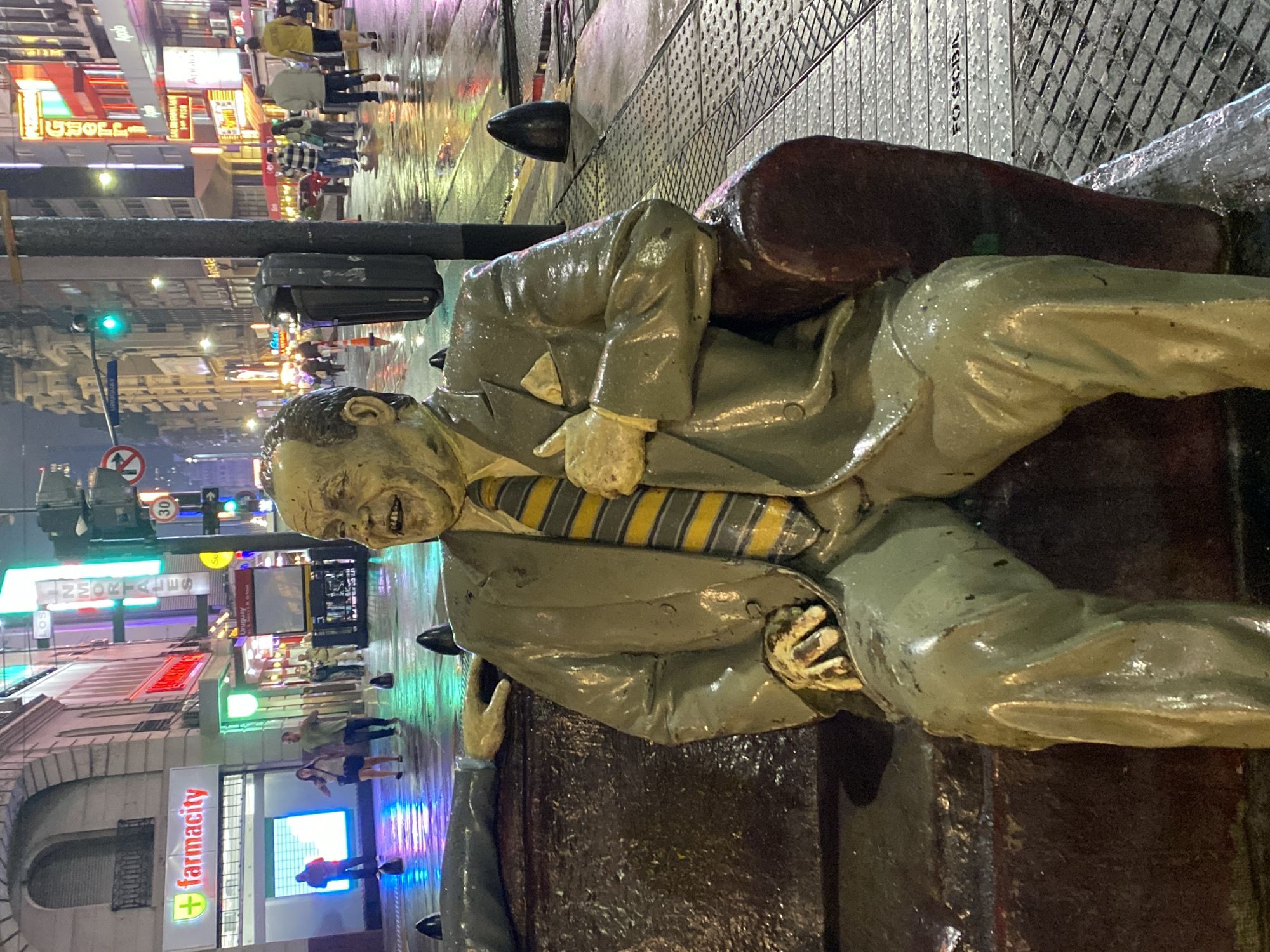 The image displays a nighttime urban street scene on Corrientes Avenue, near Uruguay Street, in Buenos Aires, Argentina, characterized by wet surfaces reflecting artificial lights. The primary subject is a life-sized, weathered statue of Alberto Olmedo, a prominent Argentine comedian, seated on a reddish-brown bench in the immediate foreground. Olmedo is depicted with a wide, open-mouthed smile, wearing a greenish-grey suit, a yellow and grey striped tie, and a white pocket square. His right hand rests on his lap, while his left hand is raised to his chest with the thumb pointing upwards. A partial view of another statue's arm is visible to the left, resting on the same bench.
The background features a bustling commercial street with multi-story buildings. Prominent illuminated signs include "Farmacity" (a pharmacy with a red and green logo) and "LOS INMORTALES" (a pizzeria with a tall, brightly lit sign). Numerous other neon signs in red, blue, and green hues adorn the buildings. The sidewalks and street are wet, reflecting the vibrant lights. Pedestrians are scattered across the sidewalks, walking in various directions. Street furniture includes a black lamppost with an attached waste bin, traffic lights (one showing green), and a "No U-turn" sign. A subway station entrance sign for "Uruguay" is also visible. The sidewalk incorporates sections of a textured metal grate, partially inscribed with "FO GOBA."