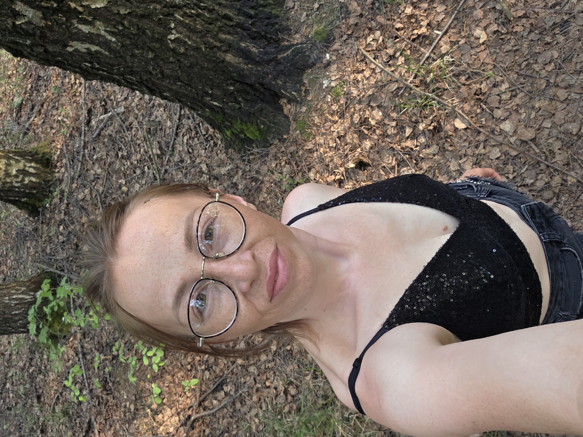 A woman with light skin and light brown hair is wearing black-rimmed glasses, a black sequined top, and dark jeans in an outdoor, wooded area. The ground is covered in brown leaves and twigs. A tree is visible to the left of the woman. The location appears to be Chaikovsky, Russia.