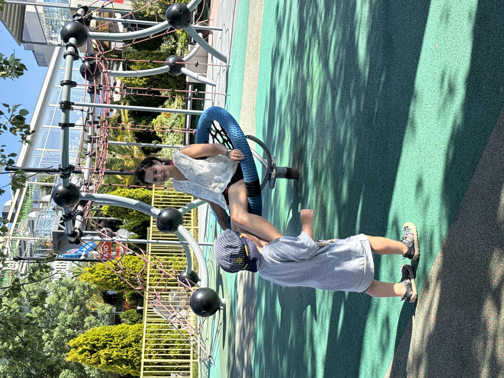 The image captures a bright, sunny day at a children's playground in Sochi, Russia. In the foreground, a young boy wearing a grey t-shirt and a striped baseball cap faces away from the camera, his body angled towards a woman seated on a blue circular swing. The woman, dressed in a white sleeveless top and dark shorts, is looking towards the boy, her left leg extended. The playground features a soft, green, impact-absorbing surface with a contrasting grey border in the foreground. In the background, a complex climbing structure with black spherical elements and netting is visible, along with lush green trees and a modern building with glass windows. The lighting suggests it is midday, with distinct shadows cast on the playground surface.