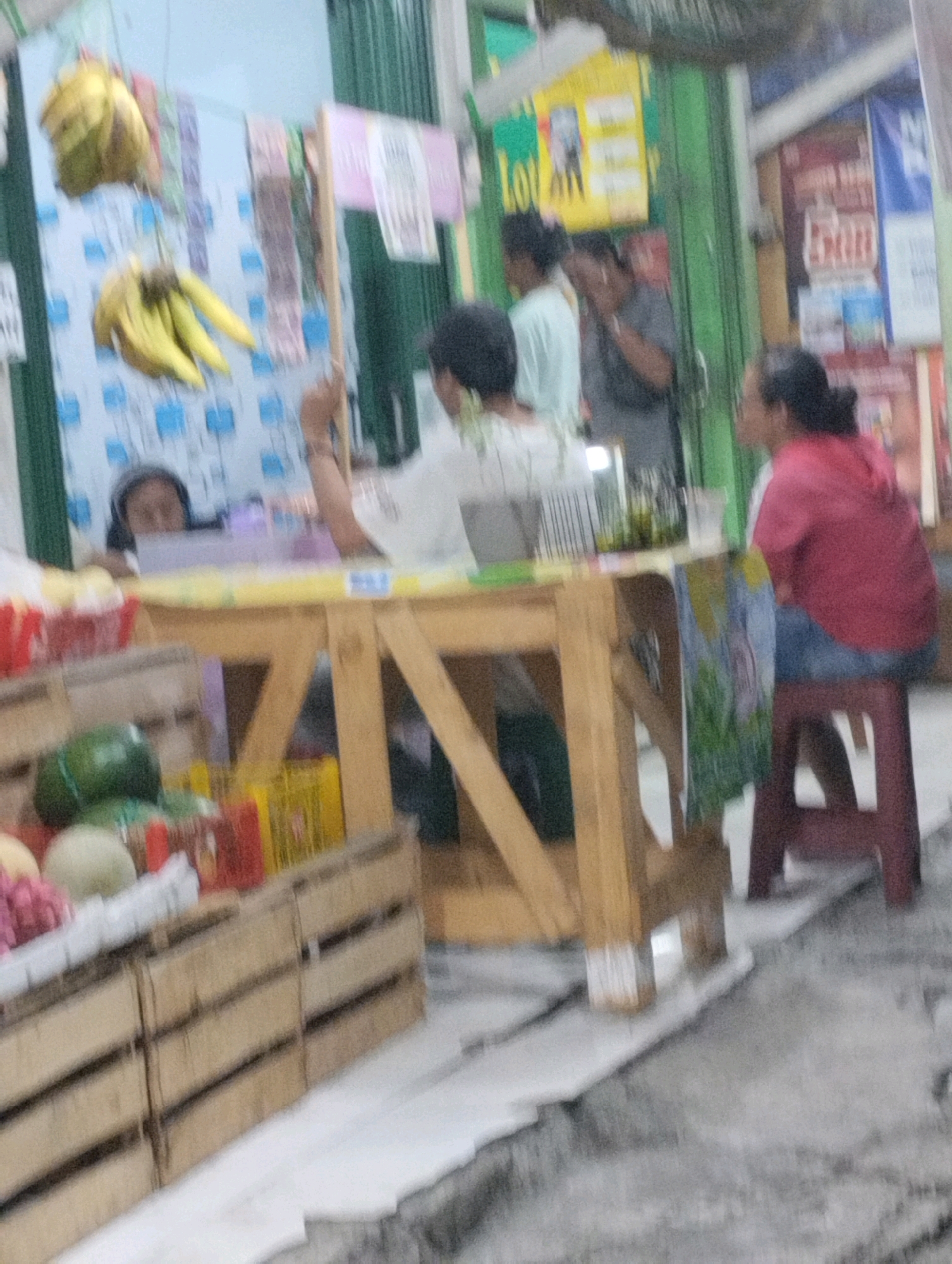 An outdoor market stall in Bekasi, Indonesia, displays various fruits and vegetables. A young girl with dark hair, wearing a light-colored shirt, is seen behind the counter, looking down. To her right, a woman in a red hoodie and blue jeans is seated on a maroon stool. Behind the stall, several people are visible, some with dark hair, engaged in what appears to be casual conversation or work. The stall itself is made of light-colored wood, and a variety of produce, including bananas, watermelons, and other fruits, are neatly arranged in wooden crates and on display. The background shows the typical elements of a market, with posters and awnings suggesting a bustling environment. The overall scene captures a moment in the daily life of a local marketplace.