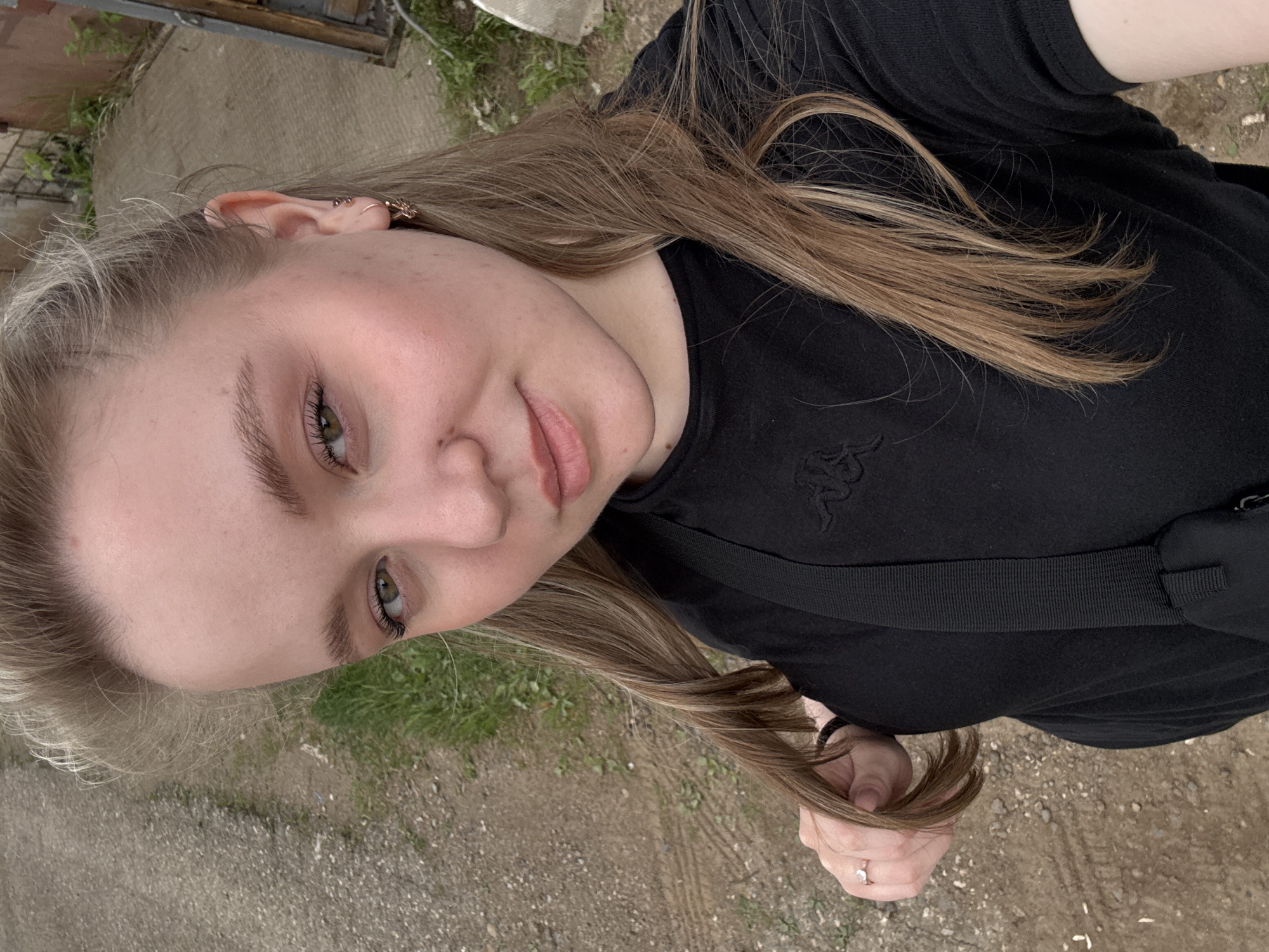 A young woman with light brown hair and fair skin poses for a selfie outdoors. She is wearing a black t-shirt with a visible Kappa logo and a black crossbody bag. Her hair is styled with lighter highlights. She is wearing earrings in her left ear and a ring on her right hand. The background consists of a dirt or gravel path with patches of grass and greenery. A partially visible grey structure or door is located in the upper left corner. The location appears to be an urban or semi-urban area, possibly a park or residential neighborhood in Chaikovsky, Russia.