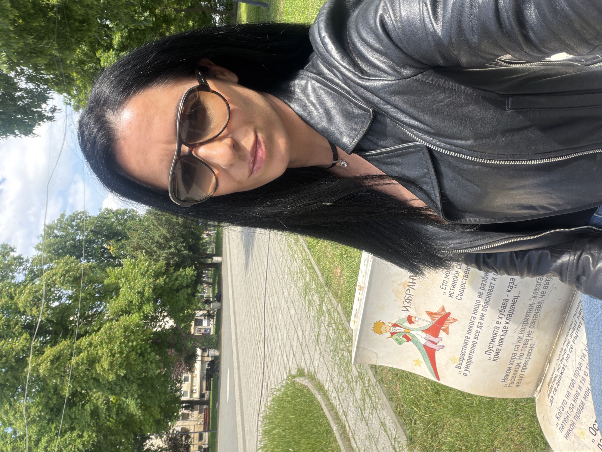 A woman with long, dark hair, sunglasses, and a black leather jacket is posing for a photo on a sunny day. She appears to be sitting on a park bench. The bench has an illustration and text from "The Little Prince" by Antoine de Saint-Exupéry in Bulgarian. Cobblestone paving and trees are visible in the background, suggesting a park setting. Based on the image content, the location is in Pleven, Bulgaria, as the "The Little Prince" themed bench is part of a series of literary benches in the city.