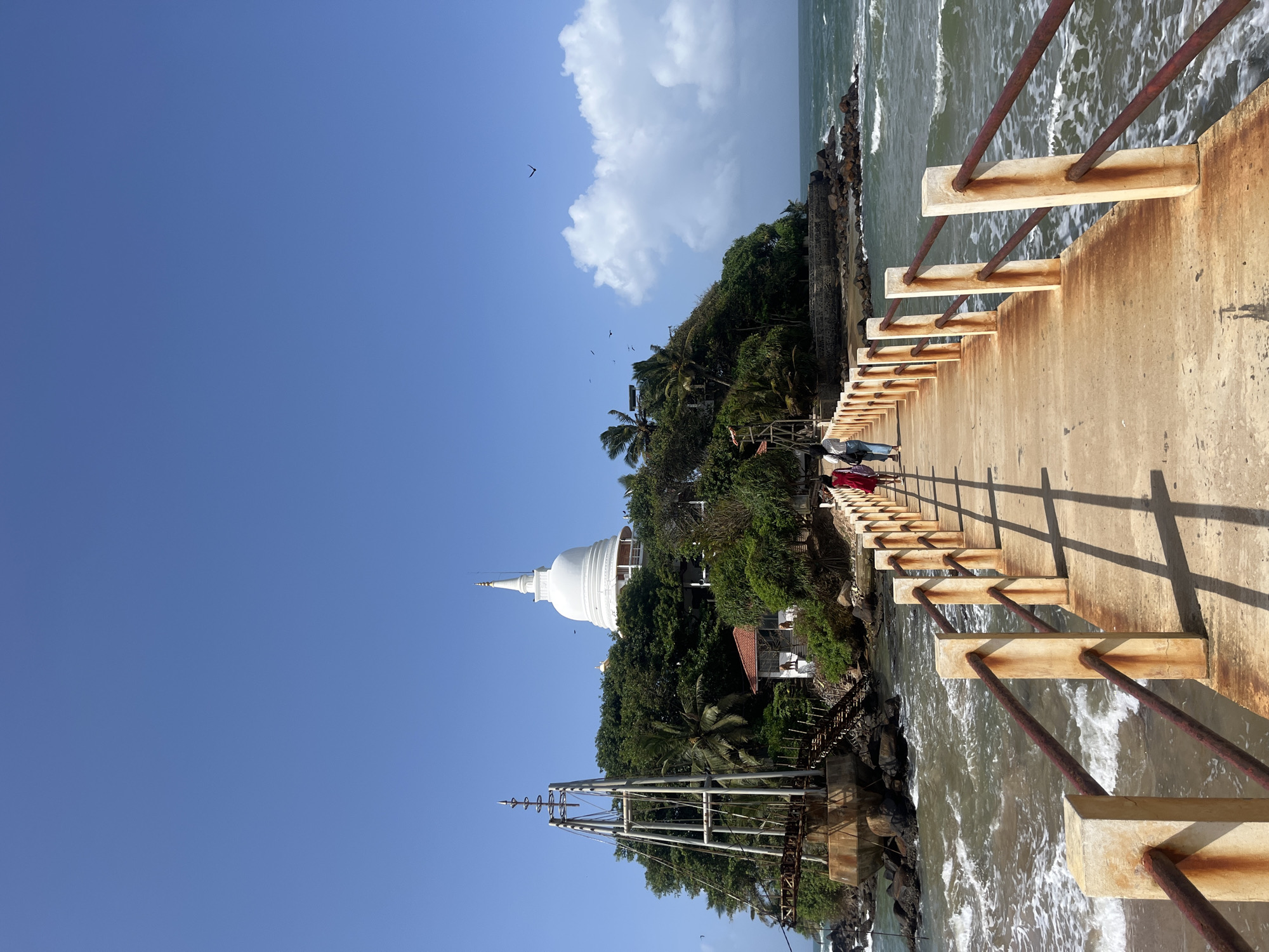The image depicts a bridge leading to an island with a white stupa in Walgama, Sri Lanka. The bridge is made of concrete with rusty metal railings. Two women are walking on the bridge towards the island. The island is covered in lush green trees, and a white stupa with a spire is visible at the top. The sea surrounds the island, with waves crashing against the shore. In the distance, a large white cloud is visible in the blue sky, and several birds are flying. A rusty metal structure, possibly part of an old bridge or pier, is located on the left side of the island. The overall scene is serene and picturesque, showcasing a blend of natural beauty and religious architecture.
