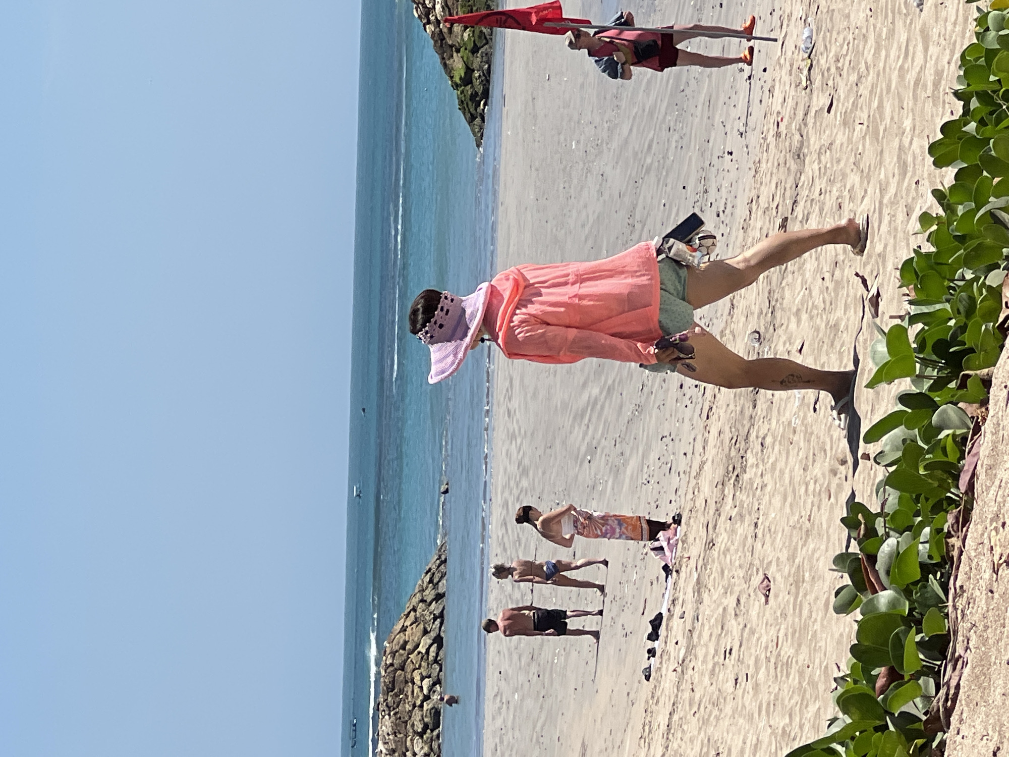 The image depicts a beach scene in Kuta, Indonesia, under a clear blue sky. In the foreground, green vegetation lines the bottom edge of the frame. The sand is light brown and appears somewhat littered.
Several people are visible on the beach. A person in the mid-ground, facing away from the camera, wears a pink sun visor and a bright pink hooded jacket over light green shorts. They are holding sunglasses and a dark object in their right hand. To the left of this person, three individuals stand on the sand: a shirtless man wearing dark shorts, a woman in a bikini, and another woman wearing a white top and an orange and pink sarong. Further to the left, another person is partially submerged in the ocean near a stone breakwater.
In the background, the turquoise ocean meets the horizon. Waves are visible, and several small boats are scattered on the water. Two more breakwaters, constructed from large stones and covered in green mossy growth, extend into the sea. On the right side of the image, a person in a red vest and shorts holds a red flag attached to a pole.
The overall impression is a sunny day at a popular beach destination.