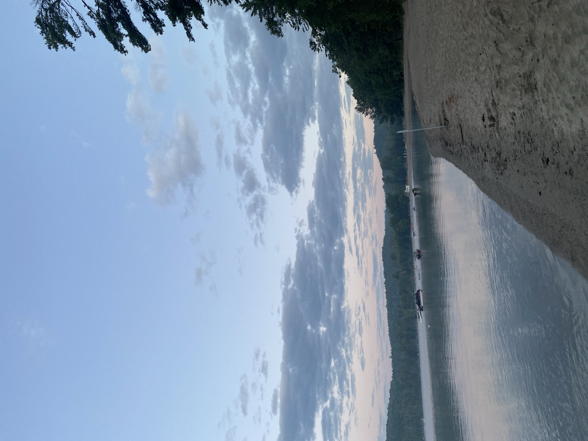 The image depicts a serene natural landscape at the shoreline of a lake in Duhamel, Canada. The sky is a pale blue with scattered clouds, tinged with pink hues near the horizon, suggesting either dawn or dusk. A gentle mist hovers over the calm water, reflecting the sky and the distant tree-covered hills. Several boats are visible on the lake, some anchored, others potentially moving.
The foreground features a sandy beach, with a distinct line where the sand meets the water. A thin white pole stands upright on the beach, likely marking a boundary or a path. The right side of the image is dominated by a dense line of dark green coniferous trees, which extend upwards and frame the sky. The overall atmosphere is tranquil and peaceful, characteristic of a natural lakeside environment.