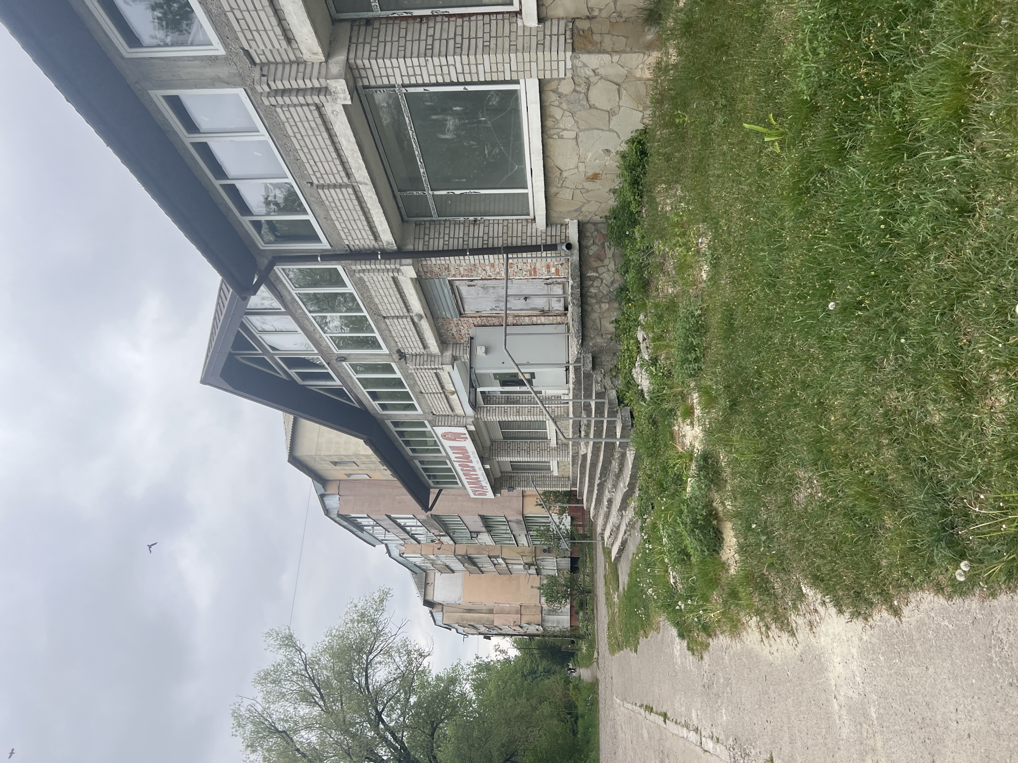 The image depicts a street scene in Cherliany, Ukraine. Buildings line one side of a paved walkway. The structures vary in architectural style, including multi-story apartment buildings and a brick-faced building with a large window. The brick building has signage in Russian that reads "БУХГАЛТЕРСКИЙ" The walkway is bordered by grass. A bird flies in the grey sky. Trees with green foliage are visible in the background.