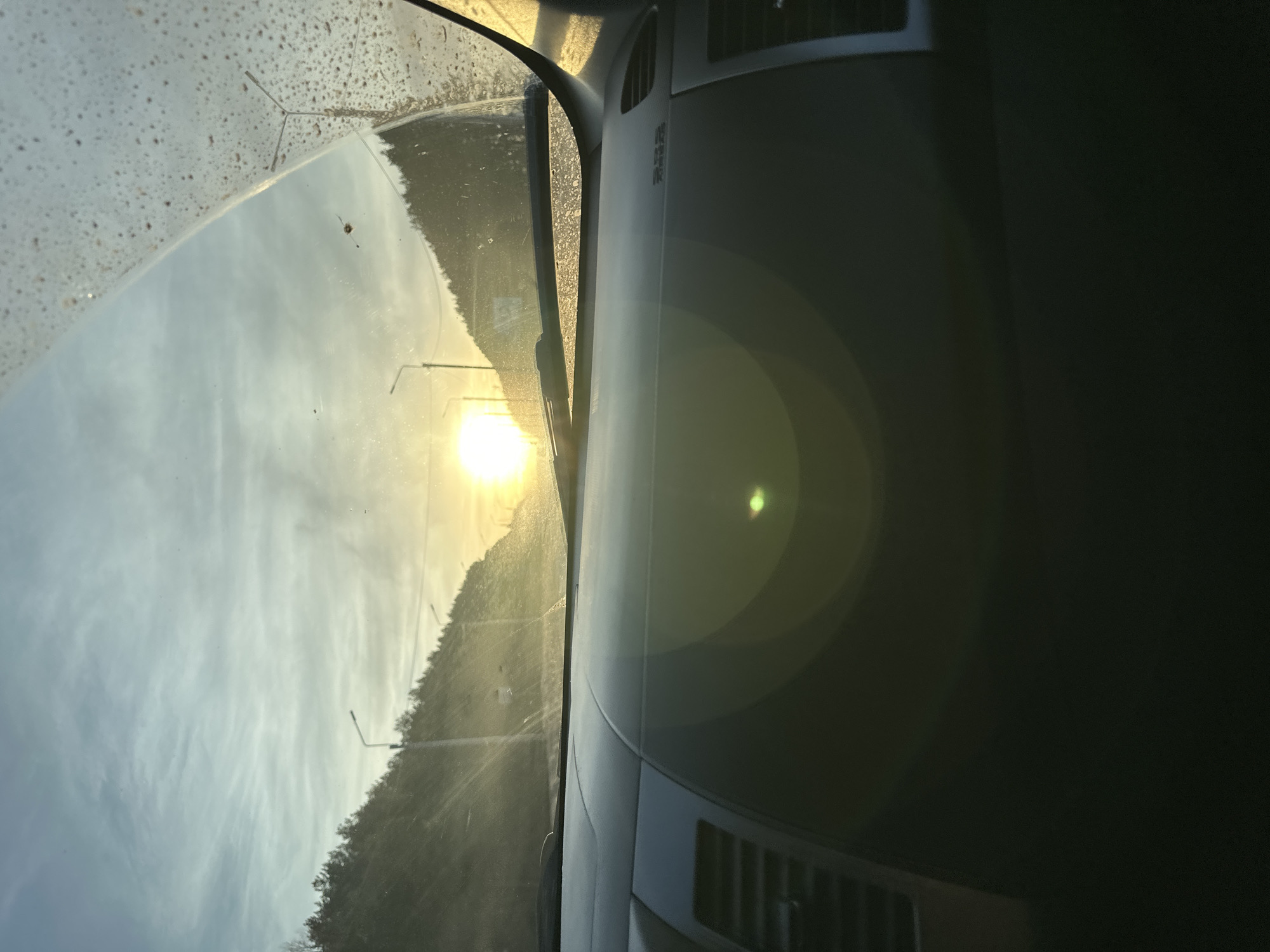 The image displays a road scene in or near Shud'ya, Russia, captured from the interior of a vehicle, likely during sunrise or sunset. The lower half of the frame is occupied by a dark grey vehicle dashboard, featuring an air vent on the far left and a horizontal windshield wiper blade visible at the base of the windshield. The windshield exhibits numerous small dirt particles and water spots, predominantly on the upper right quadrant. A bright, intense yellow-orange sun is positioned low on the horizon, centrally within the upper half of the frame, creating significant glare and a green lens flare reflected on the dashboard. The sky above the horizon is light blue with sparse, thin clouds. The implied road ahead is flanked by two steep hills, densely covered with dark green coniferous trees. Multiple tall, slender streetlights with single fixtures are visible along the unseen roadside, extending towards the sun's position. No individuals are visible in the scene.