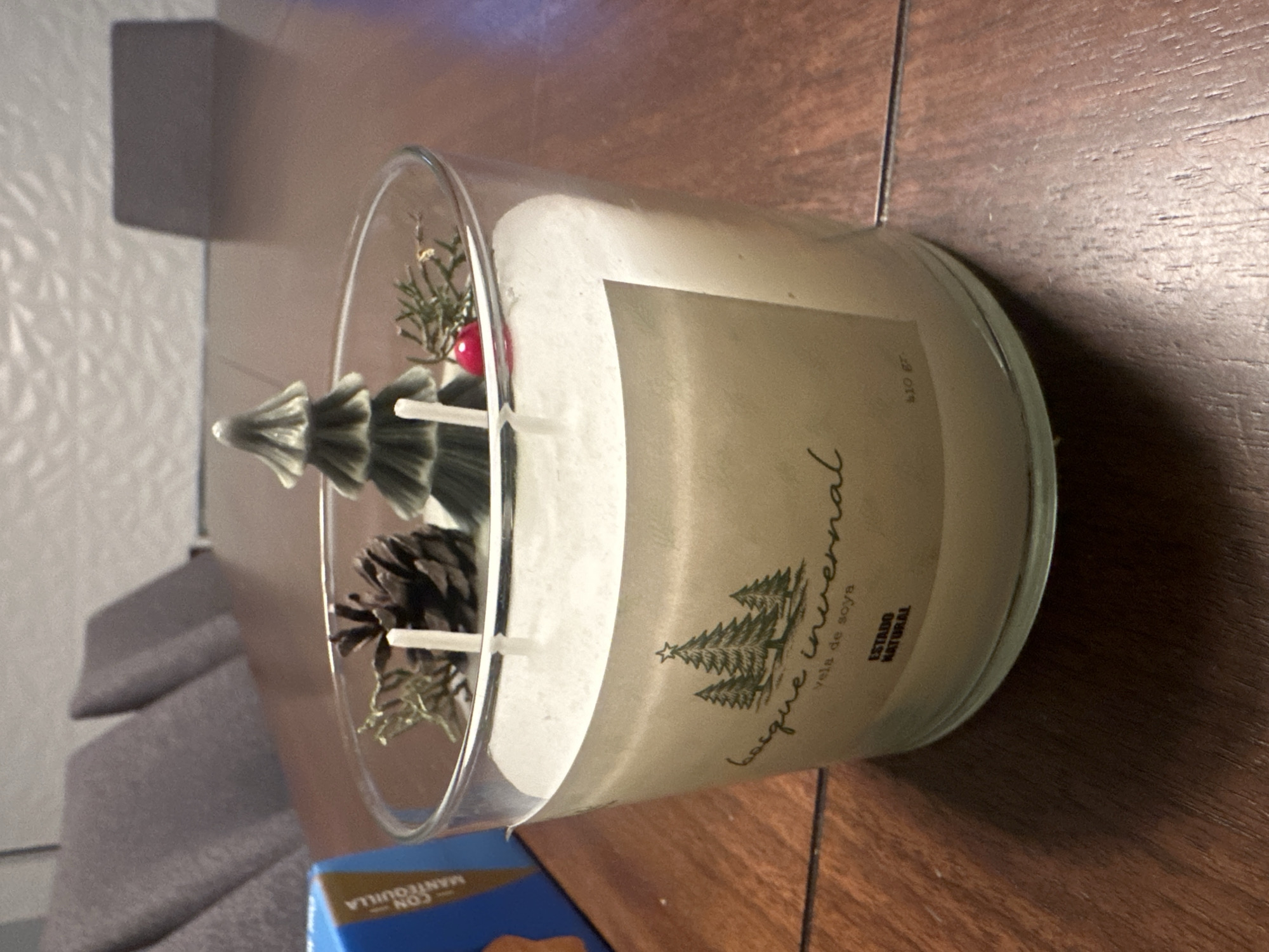 A clear glass jar candle sits on a wooden table, with a blue box to its left and chairs in the background. The candle is filled with white wax and features two wicks. Decorative elements, including a pinecone, a small green Christmas tree figurine, and a red berry with sprigs of evergreen, are embedded in the top layer of the wax. A label on the front of the candle reads "Bosque Invernal vela de soya ESTADO NATURAL 410 gr." The scene is indoors, likely in a residential setting. There are no people or animals visible. The lighting suggests an indoor artificial light source.