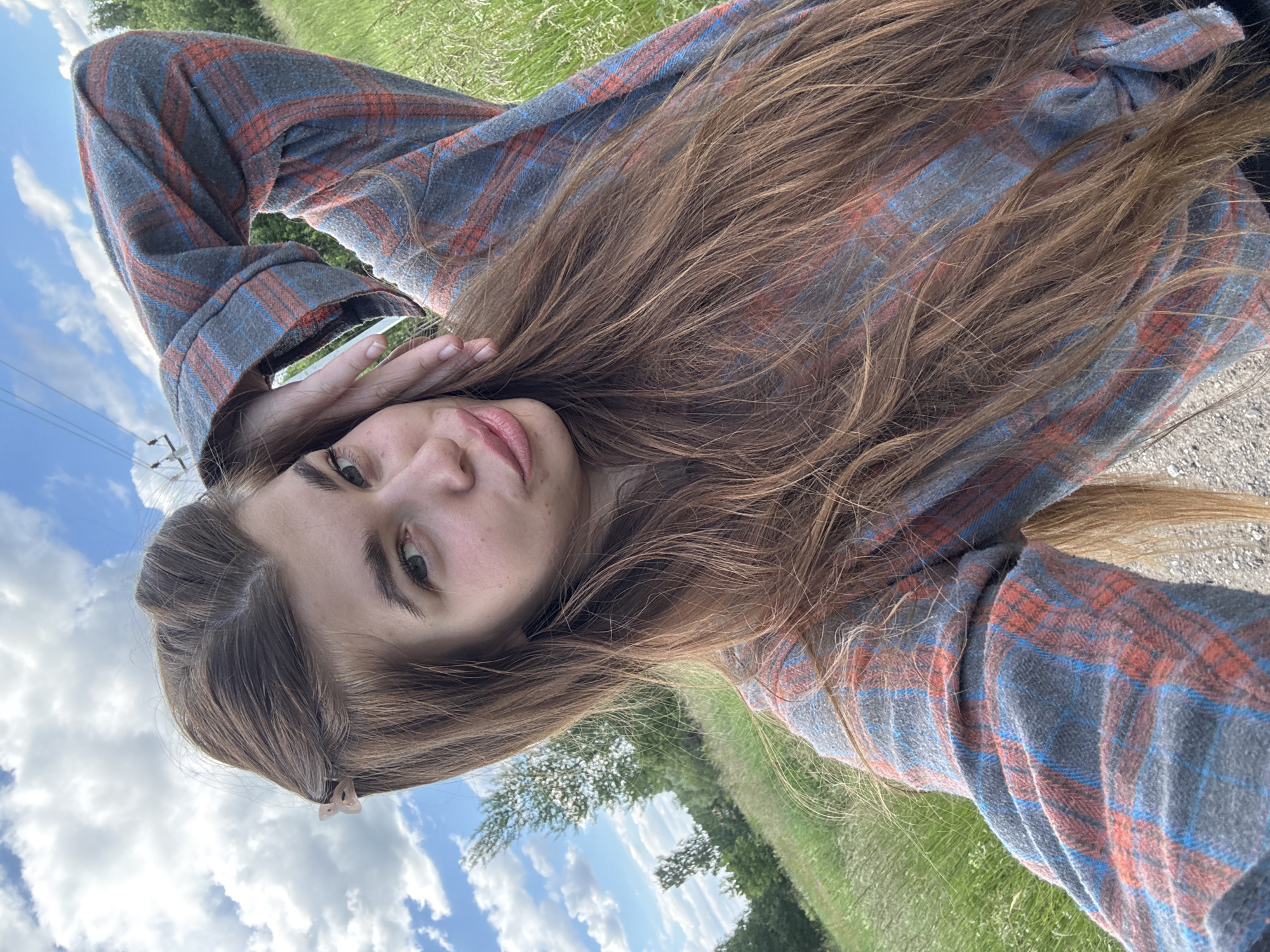 The media file shows a young woman lying on her back in a grassy field under a cloudy blue sky. She is wearing a plaid shirt with blue and red stripes. Her long, brown hair is spread out around her. Her right hand is raised to her face, partially obscuring her left eye. Her lips are slightly parted, and she appears to be looking at the camera with a relaxed expression. The background consists of a field of green grass, some trees in the distance, and a bright blue sky with fluffy white clouds. The overall impression is one of a casual and peaceful moment outdoors. The location is identified as Meshchera, Russia. The time of day appears to be daytime, given the bright sky. There is no visible text in the media file.