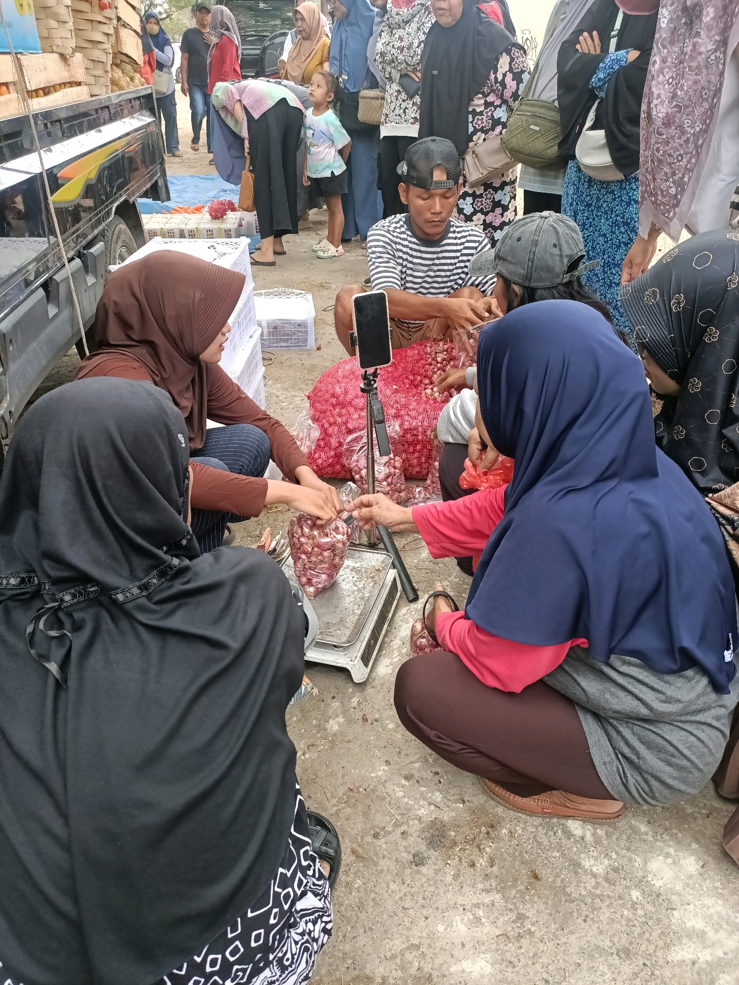 At a street market in Madiun, Indonesia, a group of people are gathered around a scale. The foreground features two women wearing hijabs, one in black and the other in brown. The woman in black is seated, facing away from the camera, while the woman in brown is also seated, facing towards a pile of red onions. She is wearing a brown hijab and a long-sleeved shirt. To her right, a third woman in a blue hijab and pink shirt is also crouched down, interacting with the onions. A man in a striped shirt and a baseball cap is seated behind a pile of onions and is looking down at the scale. He is wearing a black cap. In the background, a truck is parked, and more people are visible, including a young child in a colorful t-shirt standing and observing. Crates and large bags of produce, likely onions, are scattered around the scene. The ground appears to be concrete or asphalt. A tripod with a phone is set up to the right of the scale, suggesting the activity might be recorded.