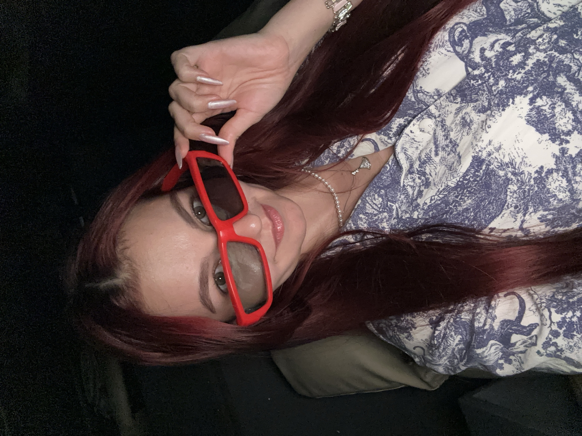 A woman with long, dark red hair is taking a selfie, wearing red 3D glasses and a blue and white patterned shirt. She is holding the glasses up with her right hand, revealing long, light-colored fingernails. A pearl necklace with a small pendant is visible around her neck, as is a bracelet on her right wrist. The background is dark and appears to be indoors, possibly a living room. The woman is looking directly at the camera with a slight smile. There are no identifiable landmarks or contextual clues to suggest a location in Khan Daun Penh, Cambodia.