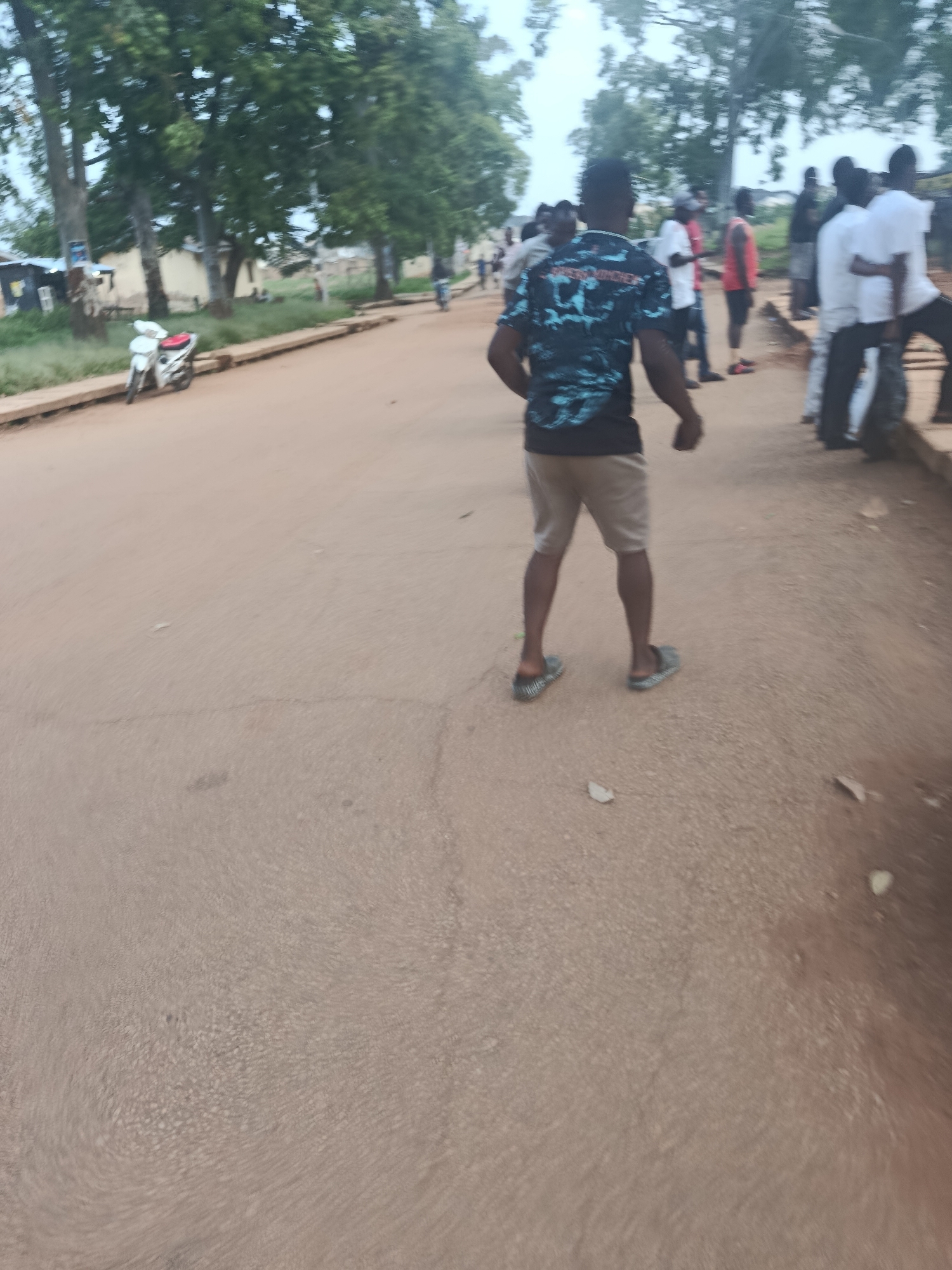 A group of people are gathered on a dirt road in Rafin Par, Nigeria. A young man in the foreground is wearing a blue and black patterned t-shirt and light brown shorts, facing away from the camera. He is standing on a dirt road with a motorcycle parked on the left side of the road. In the background, other individuals are standing and walking, some in white shirts and others in casual attire. The environment appears to be an outdoor setting with trees and buildings visible in the distance. The overall scene suggests a casual gathering or street activity.