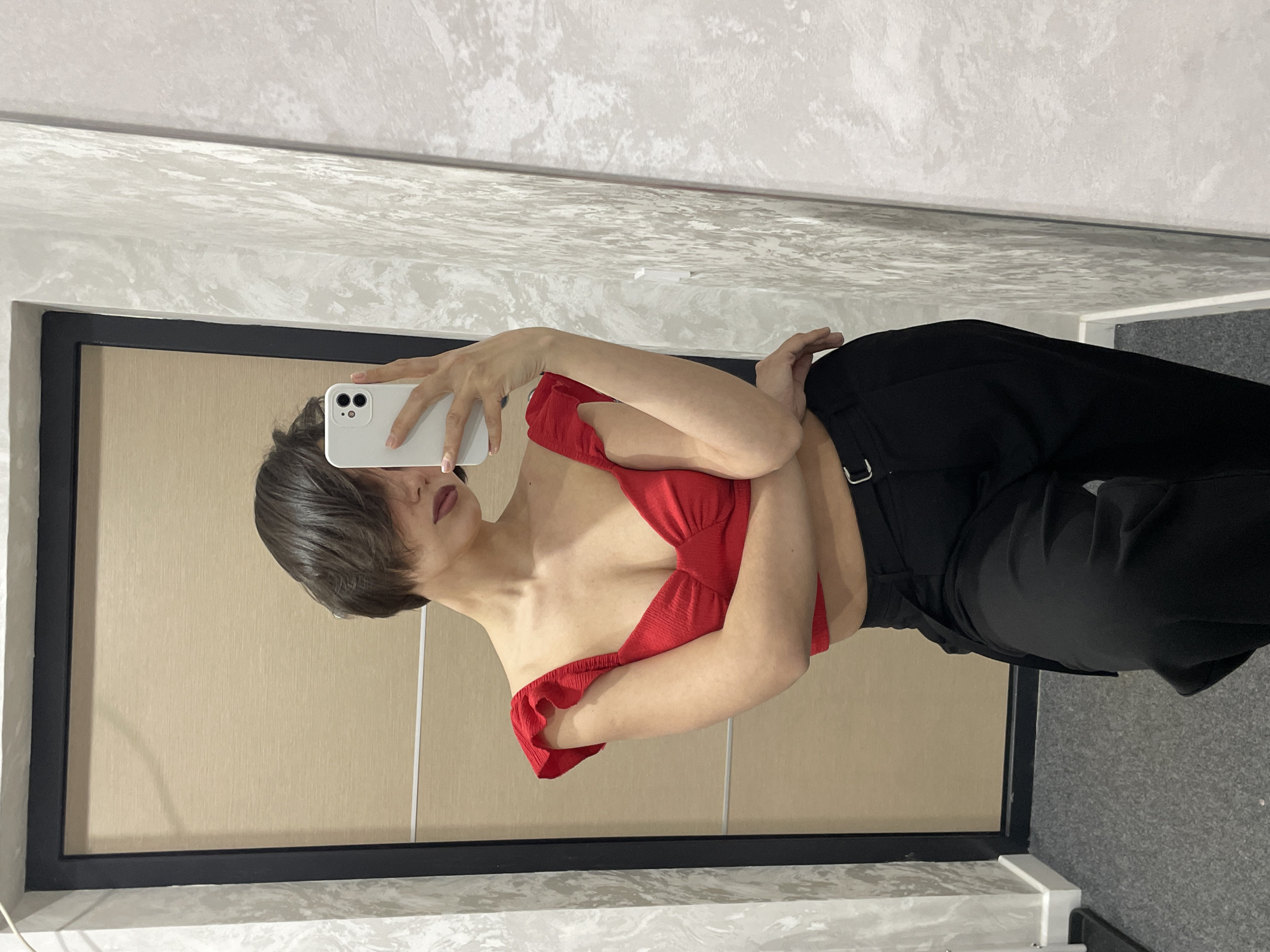 A young woman with short dark hair is taking a selfie in a mirror. She is wearing a red top with ruffled edges and black trousers. She holds a white smartphone in her right hand. The background includes a wall with a faux marble design and a light-colored panel with a black frame. The location appears to be indoors, possibly in a dressing room or bathroom. There is no specific indication in the image confirming the location as Izhevsk, Russia.