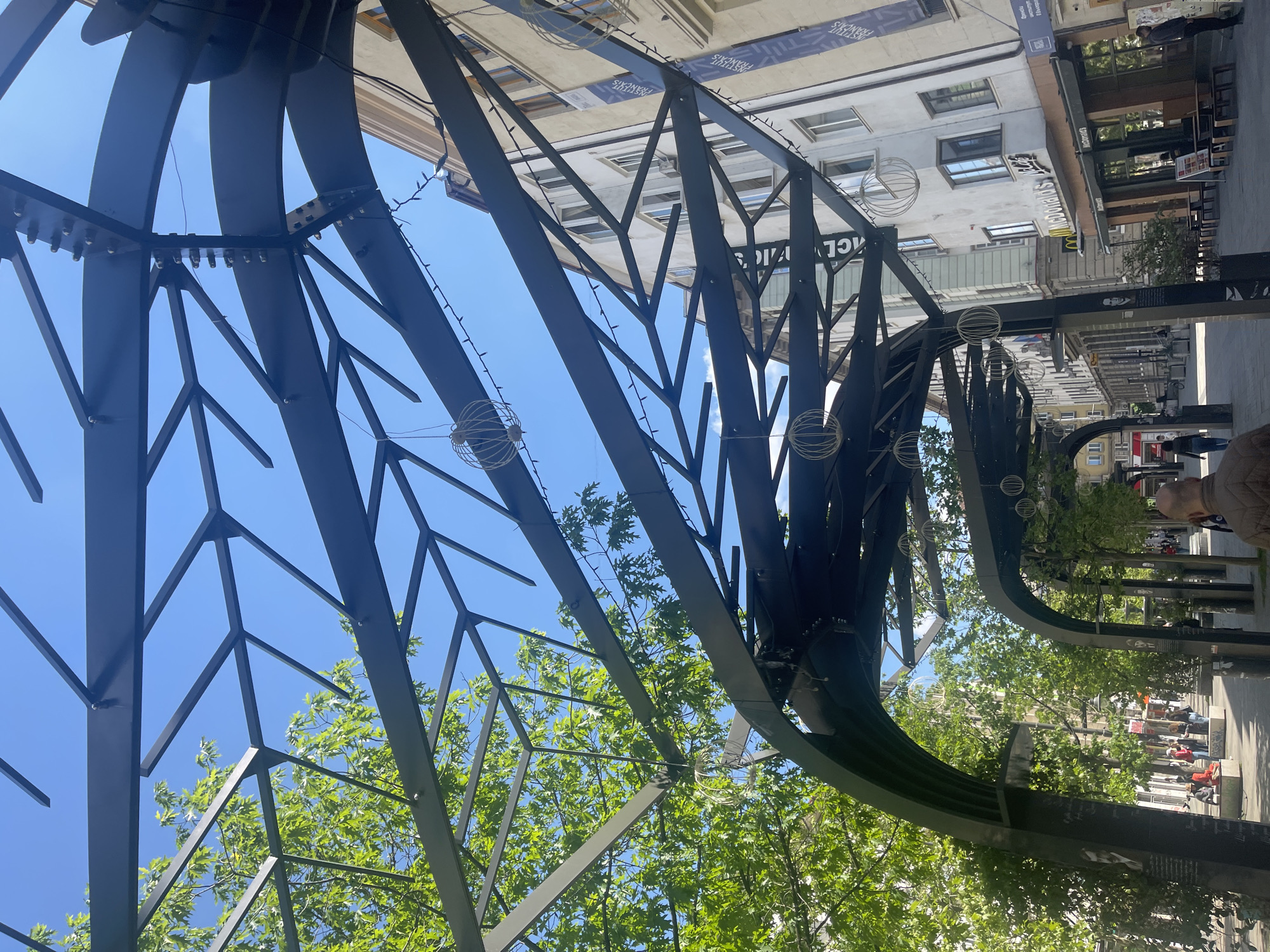 The image captures an upward view of a metallic, tree-like architectural structure in Sofia, Bulgaria. The structure, painted in a dark color, features a network of geometric lines and branches resembling a stylized tree canopy. The blue sky is visible through the gaps in the metalwork. Decorative, spherical hanging lights are attached to the structure. Green foliage of real trees can be seen intermingling with the metal branches.
In the background, buildings and street scenes are visible. The Institut Francais banner is visible on a building. Further down the street, a McDonald's restaurant is present. People can be seen walking along the street. The image is taken from a low angle, looking upwards.