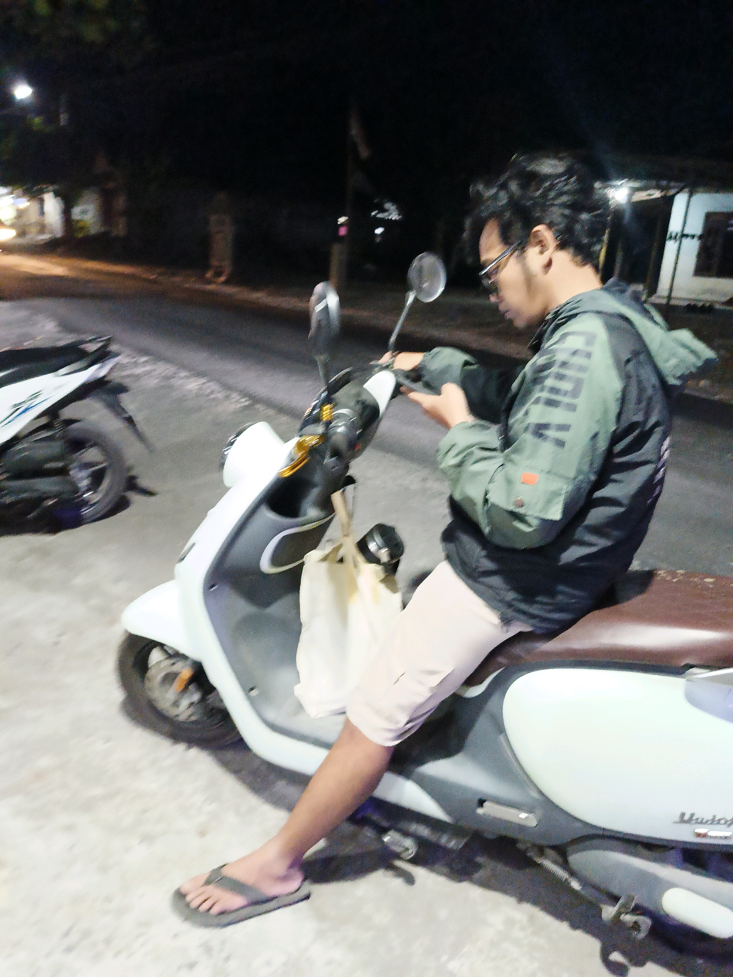 A person wearing glasses and a green jacket is sitting on a scooter at night. The scooter is light blue and white. The person is looking down at their phone. There is another scooter parked behind them. The scene appears to be a street in Manisrenggo, Indonesia, as indicated by the text on the scooter, "Mio Z", which is a Yamaha model popular in Indonesia. The background shows some lights from buildings, suggesting a residential or commercial area. The ground is paved.