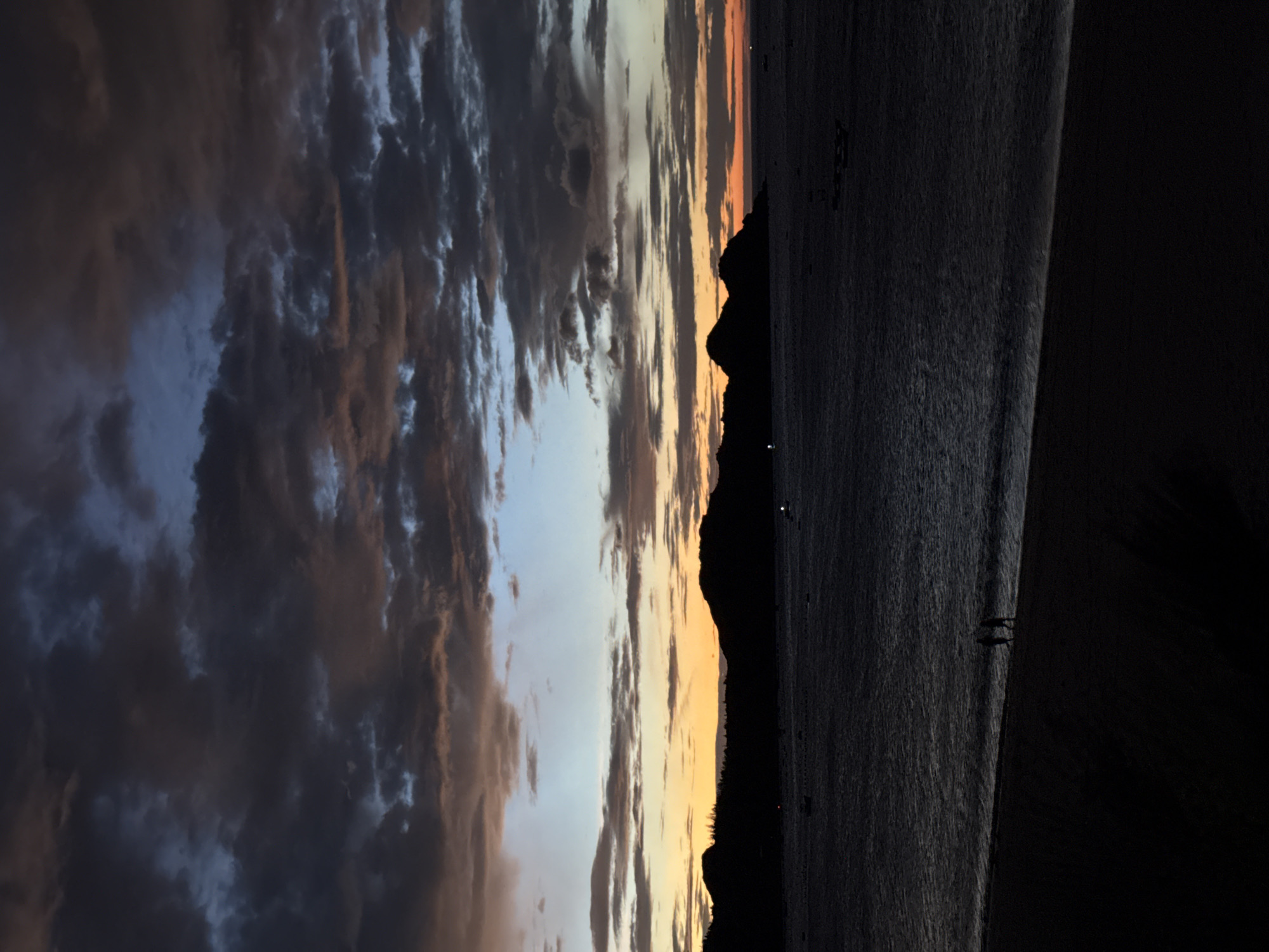 This is a vertical, low-angle photograph of a serene twilight seascape in Song Cau, Vietnam. The scene is dominated by a dramatic sky filled with layered clouds in various shades of dark blue, grey, and hints of orange and yellow near the horizon. Silhouetted against the fading light is a rugged coastline with rolling hills. The water in the foreground is dark and calm, with gentle waves lapping at a dark, sandy beach. Two small figures, silhouetted and indistinguishable in features, are walking together along the shoreline. Several small boats are scattered across the water, barely visible in the dim light. The overall atmosphere is peaceful and atmospheric, capturing the transitional moments between day and night.