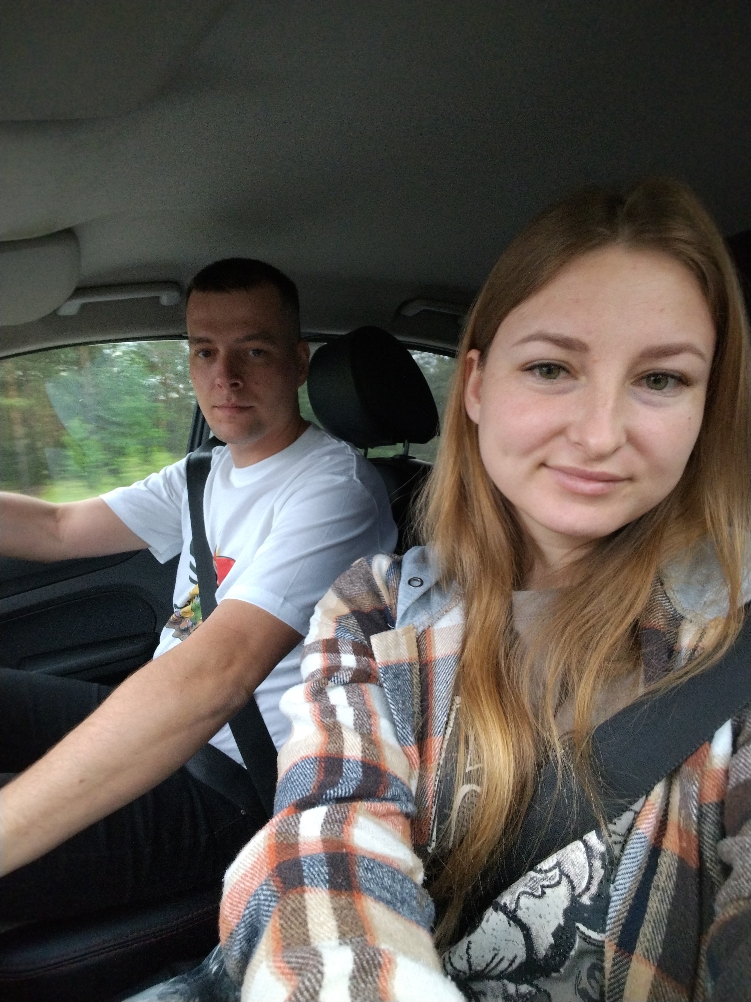 A man and a woman are seated in the front seats of a car. Both are wearing seatbelts. The man is in the driver's seat. He has short brown hair and is wearing a white t-shirt. The woman is in the passenger seat. She has long brown hair and is wearing a plaid shirt. The location is not evident from the image.