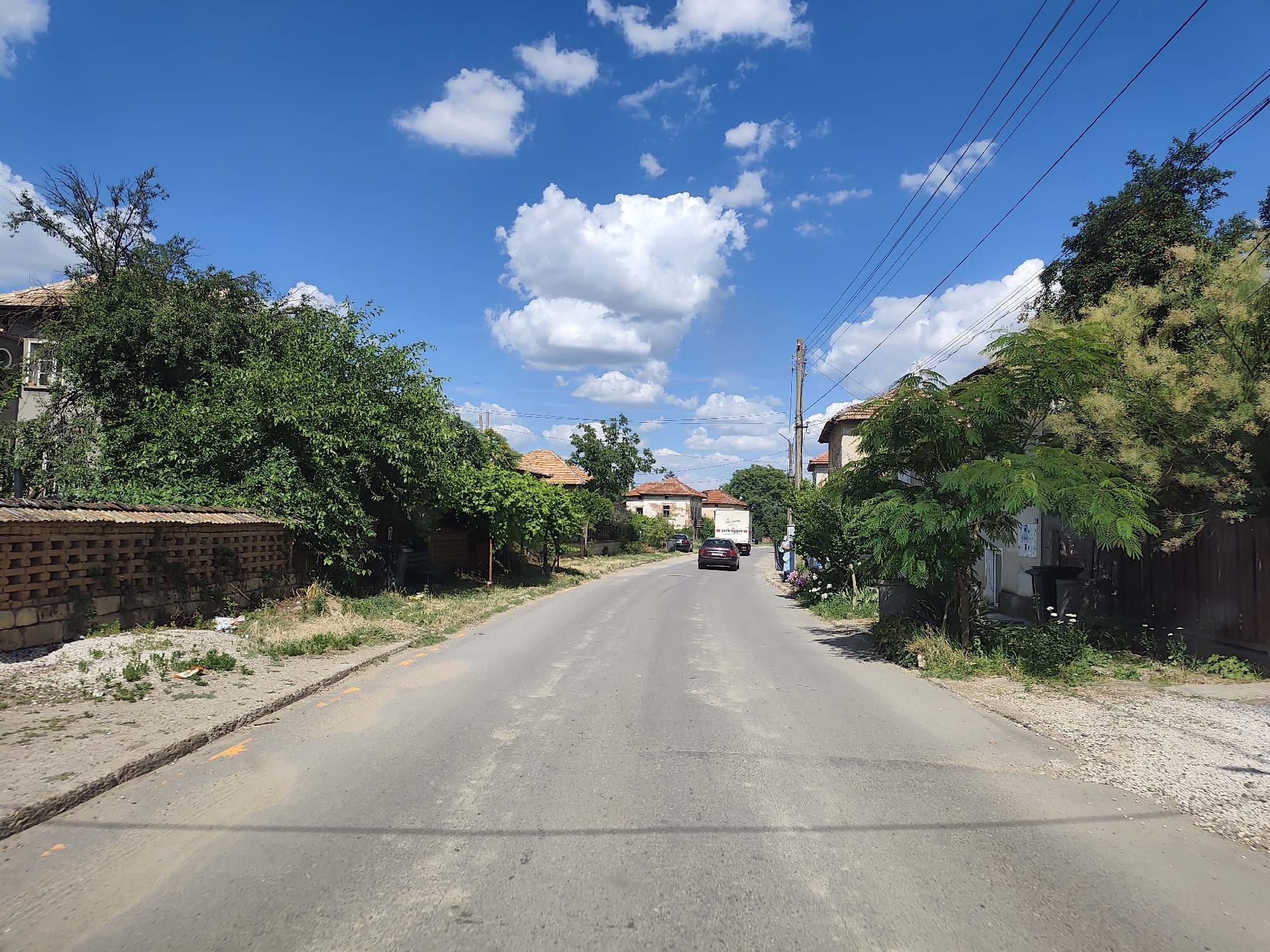 This media file depicts a sunlit, rural street scene in Sadovets, Bulgaria. The road, paved and somewhat worn, stretches into the distance, flanked by houses and dense green trees. The sky above is a vibrant blue, dotted with fluffy white cumulus clouds, suggesting a clear and pleasant day, likely midday given the strong sunlight.
On the left side of the road, a low, decorative brick wall runs alongside a property, partially obscured by lush trees. A small mound of debris is visible behind the wall. The houses on both sides of the street have traditional tiled roofs.
Further down the road, a dark-colored car is approaching, followed by a white truck with text on its side. A person can be seen standing on the right side of the road near the truck. Electrical wires crisscross the sky, supported by utility poles.
The overall impression is one of a quiet, ordinary day in a Bulgarian village, bathed in warm sunlight. No specific event or strong emotions are conveyed; the scene is peaceful and natural. No legible text is visible in the image, apart from what appears to be branding on the side of the truck.