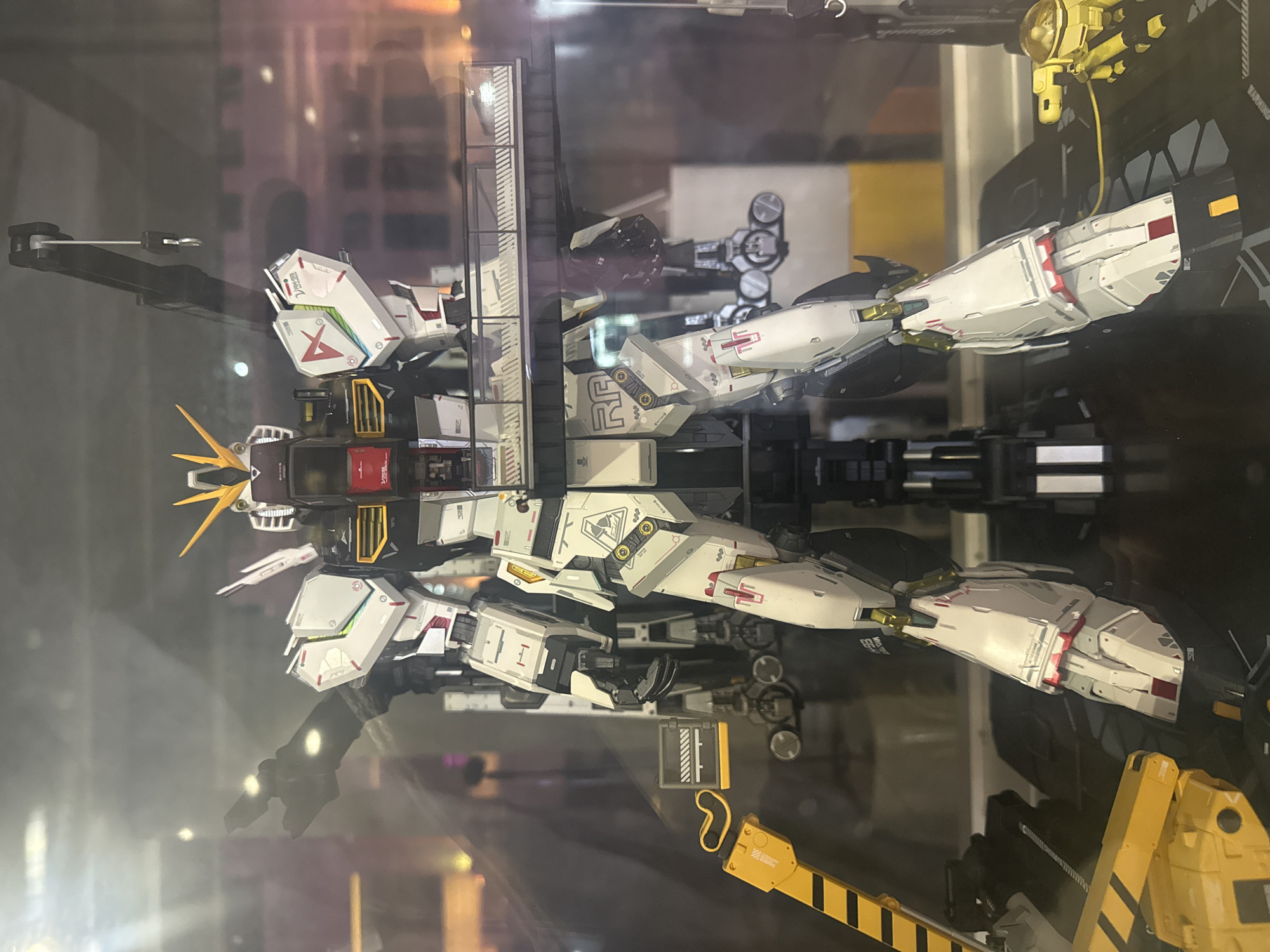 The image shows a model of a Gundam robot in a display case. The robot is primarily white with red, black, and yellow accents. It is surrounded by various structural elements, including a ladder and other machinery in yellow and black. The Gundam model appears to be part of a diorama depicting a maintenance or repair scene. The background is blurry and difficult to discern. The location is Yuexiu District, China.