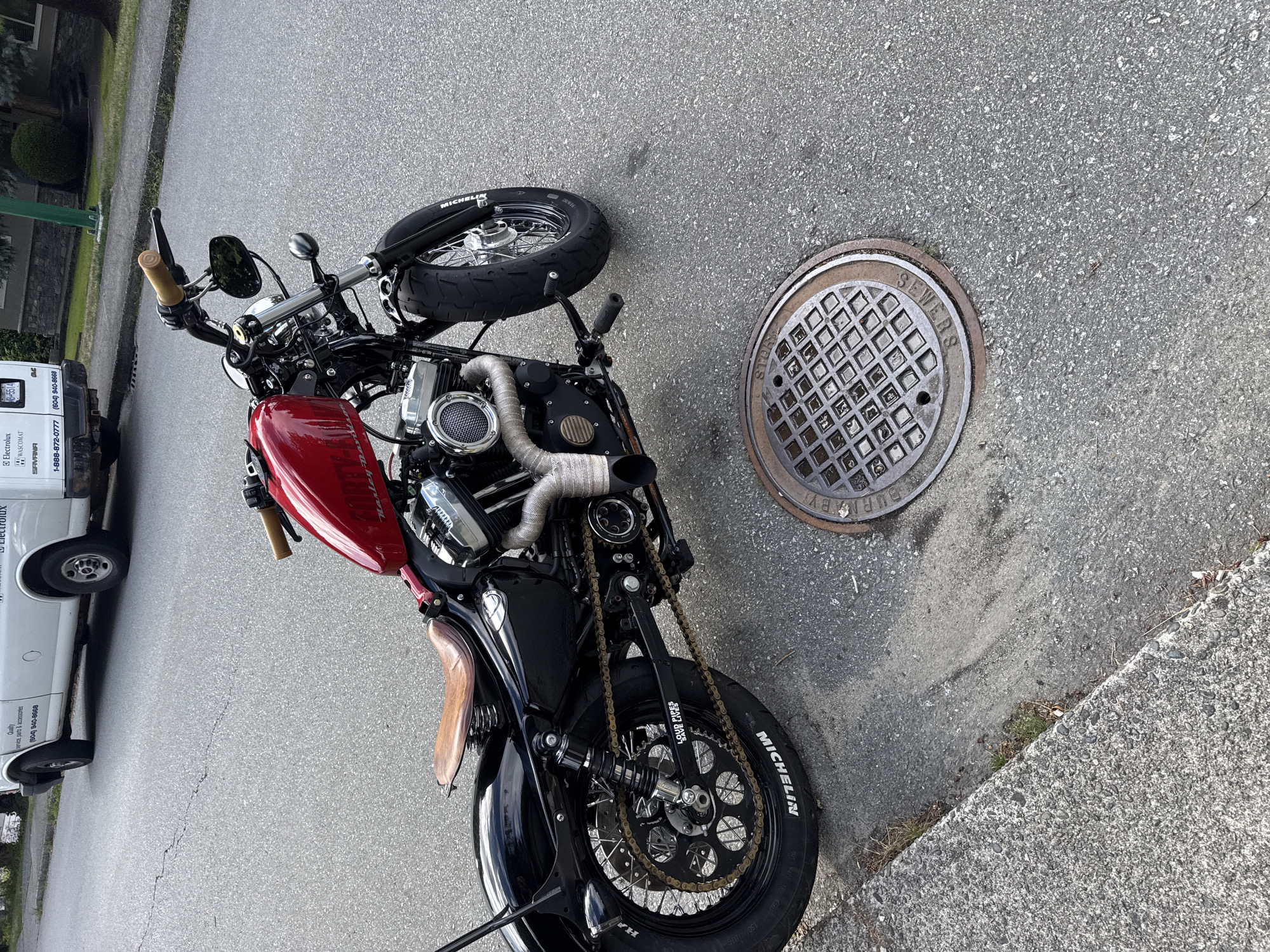 The image shows a custom red and black Harley-Davidson motorcycle parked on an asphalt street next to a concrete curb. A manhole cover is visible on the right side of the frame. The motorcycle has a brown leather seat, chrome exhaust pipes wrapped in heat tape, and a black fender with a "Loud Pipes Save Lives" sticker. The front tire is a Michelin. In the background, a white van is parked on the street. The scene appears to be a residential area in Burnaby, Canada, given the presence of a "City of Burnaby" branded manhole cover. The environment is outdoors, with trees and a building visible in the distance. The lighting suggests it is daytime, possibly overcast. There are no people visible in the image.