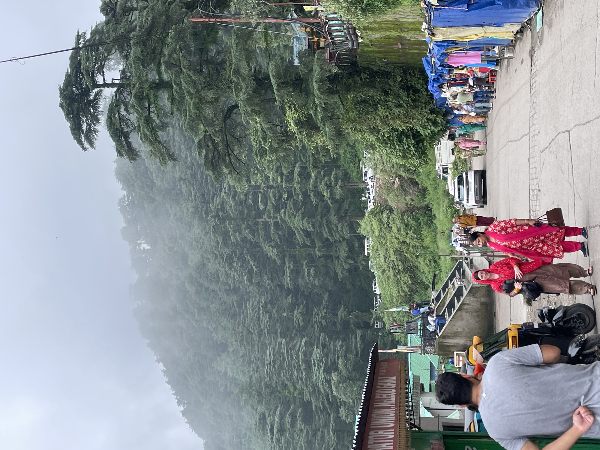 In Bhagsu, India, a road curves uphill, flanked by lush, green mountainside densely populated with cedar trees, their foliage shrouded in mist. On the left side of the road, a building with a red awning displays text that partially reads "TOR UNION, MCLEOD GANJ." A man in a grey t-shirt stands with his back to the camera, facing an auto-rickshaw. Two women dressed in traditional Indian attire walk past the auto-rickshaw. One woman wears a red outfit with a matching headscarf and carries a dark bag, while the other is in a pink dress with a floral pattern and carries a brown purse. Further up the road, several other people are walking, and vehicles, including cars and a white truck, are visible. On the right side of the road, a row of market stalls covered with blue tarps are set up, displaying various clothing items for sale. The scene depicts a bustling street in a mountainous, forested region.