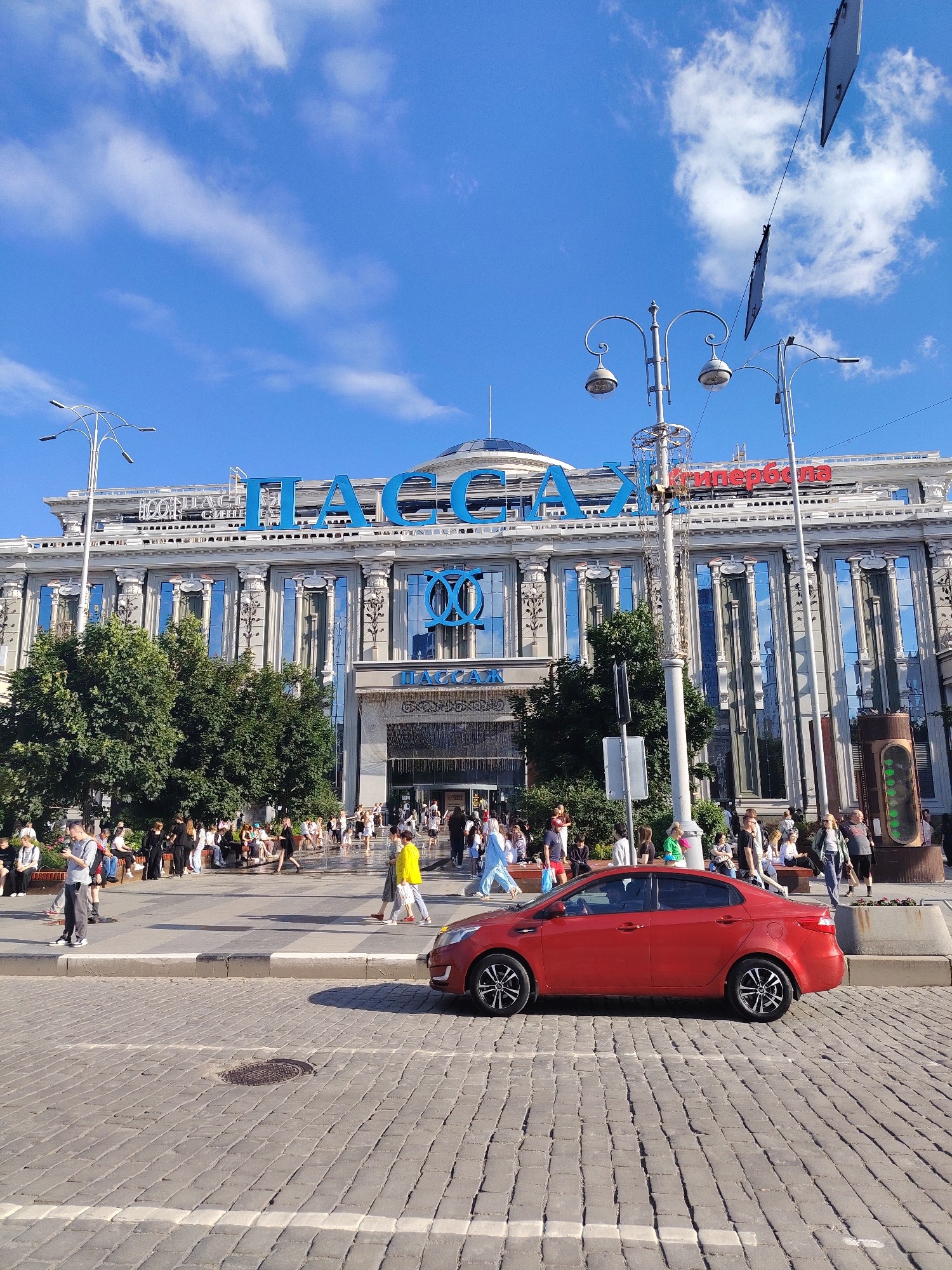 This media file shows a wide shot of a bustling city square in front of a grand, ornate building. The building, which appears to be a department store or a similar large commercial establishment, features large windows, columns, and prominent blue signage that reads "ПАССАЖ" (Passage). Red lettering above the blue sign spells out "ГУМ" (GUM), a common abbreviation for a state department store in Russia. The sky is a vibrant blue with scattered white clouds, suggesting a clear and pleasant day.
In the foreground, a red sedan is parked on a cobblestone street, facing towards the right of the frame. The street is marked with white lines, indicating a crosswalk. A circular manhole cover is visible in the cobblestones.
The midground features a wide pedestrian plaza in front of the building. Numerous people are walking, sitting on benches, and gathered in groups, indicating a lively and active scene. Some individuals are dressed in casual summer clothing. Trees are planted along the edge of the plaza, providing shade. Several ornate lampposts are scattered throughout the scene, adding to the urban aesthetic.
On the right side of the image, there's a large, somewhat unusual vertical object that resembles a traffic signal or an art installation, with a green display. The overall atmosphere appears to be daytime, likely late morning or afternoon, given the bright sunlight and shadows. The presence of many people suggests a popular public space, possibly a shopping district or a central gathering point in Yekaterinburg, Russia. There are no strong emotional cues, but the scene conveys a sense of everyday urban life and activity.