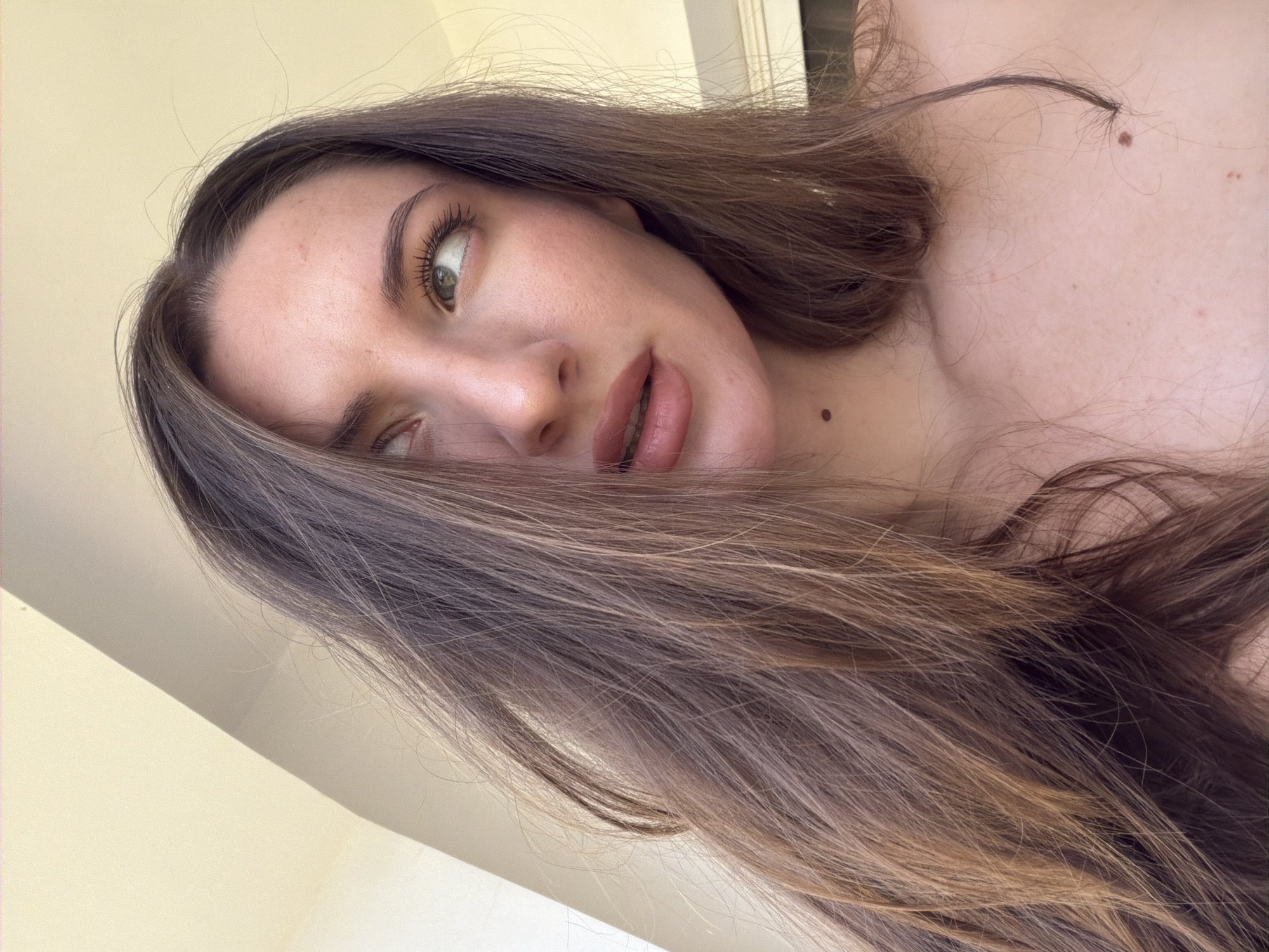 A close-up, waist-up photograph shows a young woman with long, brown, wavy hair that partially obscures her face. She is looking upwards and to her right, with her mouth slightly open. Her skin is fair, with a few freckles visible on her chest and neck. She has light green eyes and dark eyebrows. A mole is visible on her neck, just below her jawline, and another on her chest. The background is out of focus, with a light-colored wall or ceiling and a bright, possibly outdoor, light source. The image does not contain any information or context related to Sharm El Sheikh, Egypt.
