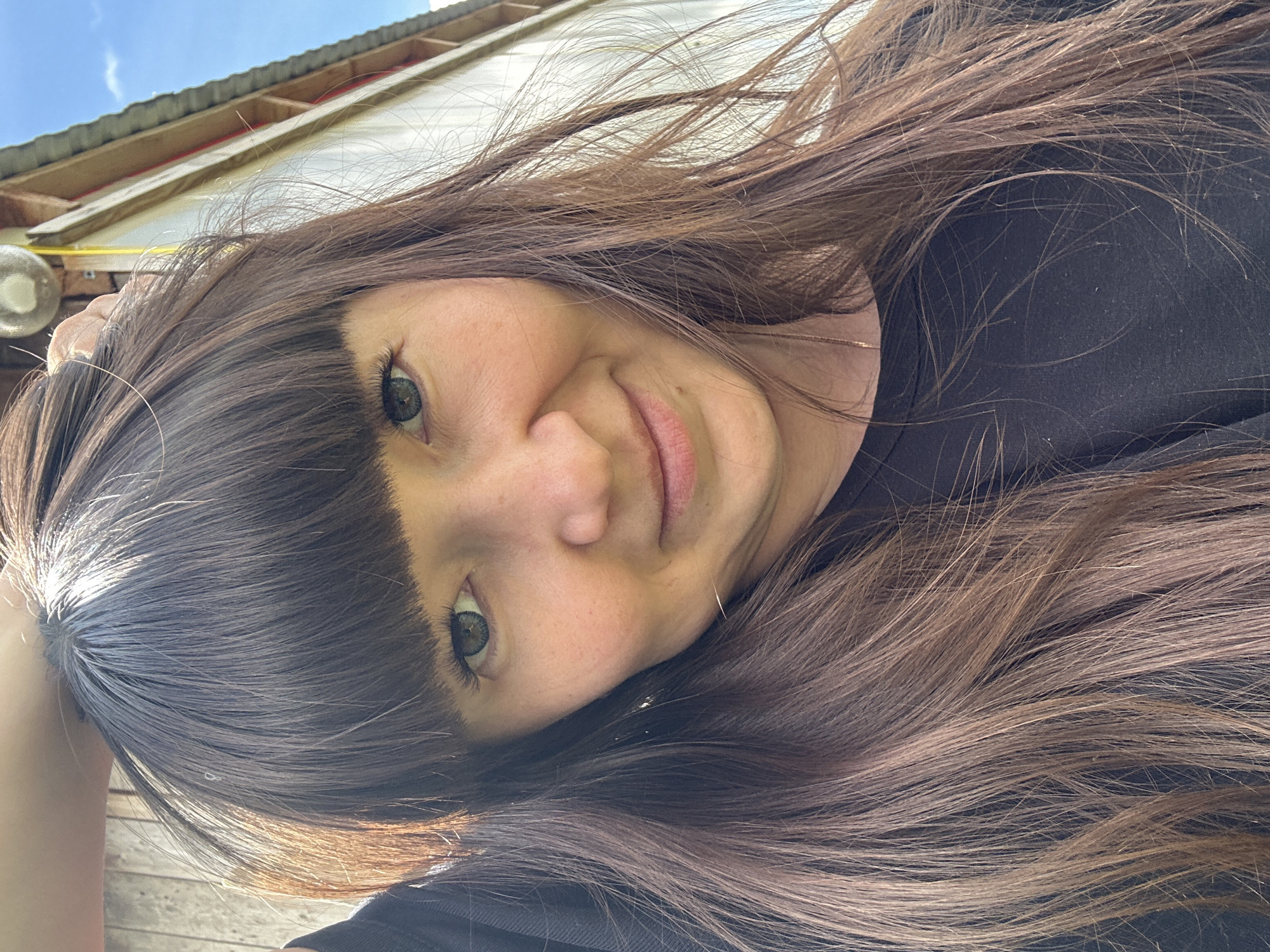 A woman with long brown hair and blunt bangs looks directly at the camera with a slight smile. She is wearing a dark shirt. The background shows part of a building with a corrugated metal roof and a light-colored wall, indicating an outdoor setting. The sky is visible in the upper left corner. The photo was likely taken outdoors in daylight. The location is Среднее Кечево, Russia.