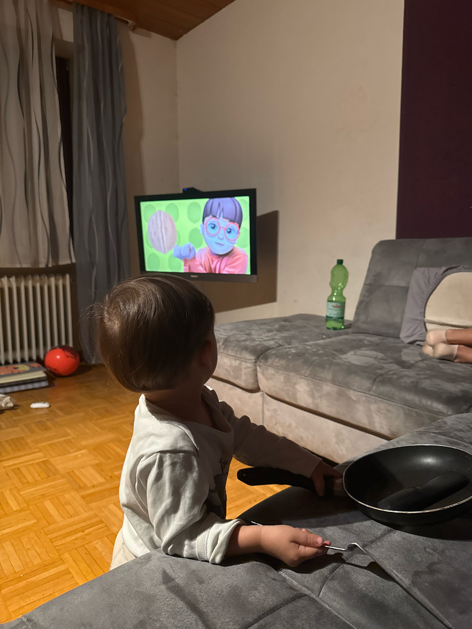 This is a medium shot of a young child in a living room, facing away from the camera and towards a television screen. The child, wearing a light-colored long-sleeved shirt and light-colored pants, is positioned on the floor in front of a dark grey sectional sofa. The child's hands are visible, with the right hand holding a fork and the left hand resting on a black frying pan situated on the sofa. The television, mounted on a wall, displays a cartoon character with glasses and a pink shirt against a green background with a walnut image.
The room features a light brown hardwood floor with a parquet pattern. To the left of the child, near a white radiator, are several stacked books and a red ball. Light grey curtains hang on the left side of the frame. On the sofa, a green plastic bottle of water is visible, and to the right, a pillow is partially obscuring another person's legs, also on the sofa. The overall lighting suggests it is evening. The scene appears to be a domestic setting, possibly in a residential area of Überherrn, Germany, given the provided location context.