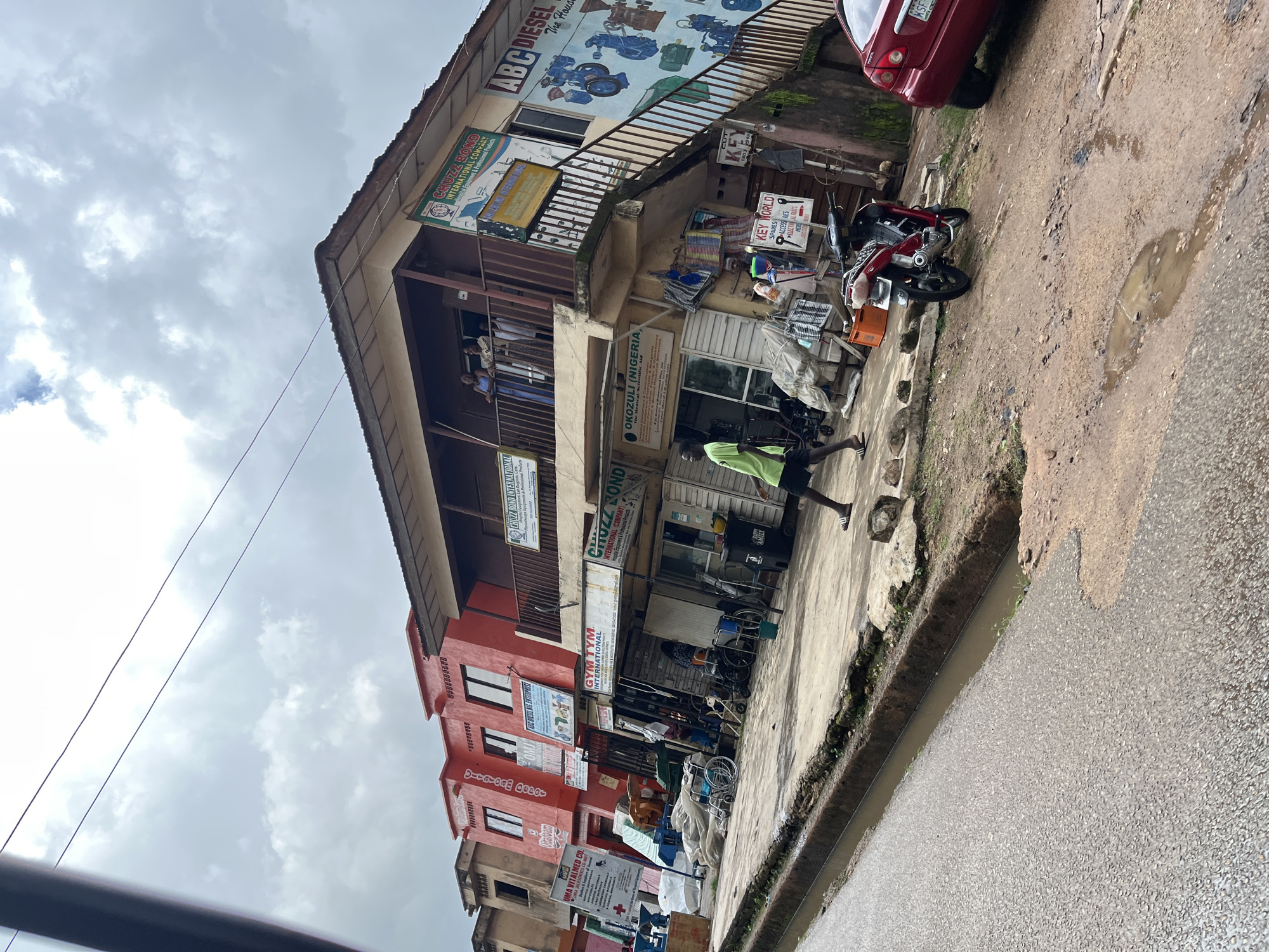 The image depicts a street scene in Jos, Nigeria, characterized by a cloudy sky and a generally overcast atmosphere, suggesting it might be daytime but not necessarily sunny. The primary focus is a multi-story building with a red lower facade and a lighter upper section, housing various businesses. Signs visible on the building indicate a diverse range of commercial activities, including "ABC DIESEL," "CHUTZ BOND INTERNATIONAL COMPANY," "OKOZULI (NIGERIA) Medical/Scientific Aid," and "GYM TYM INTERNATIONAL."
In front of the building, several objects are visible, including a red car parked to the right, and a red motorcycle. There are also several wheelchairs, some of which appear to be stored or for sale. A few people are present in the scene. One person in a bright green shirt is walking on the sidewalk, while others are standing on balconies or near the building's entrance.
The street itself is paved with a rough, textured material, and there is a water channel running alongside the sidewalk, with some standing water visible, indicative of recent rain or drainage issues. The overall impression is one of a bustling commercial area, typical of a Nigerian urban environment, with a mix of retail shops and services. The presence of multiple signs in English highlights the linguistic context.