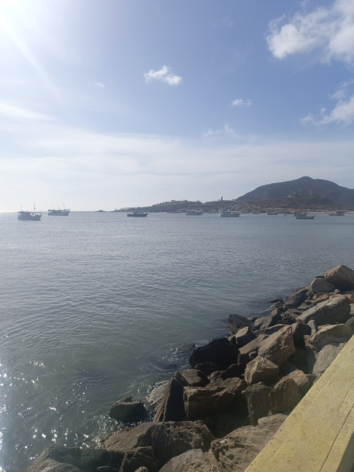 The image shows a view of the harbor in Juan Griego, Venezuela. The scene features a body of water, presumably the Caribbean Sea, with several fishing boats anchored in the distance. A rocky shoreline is visible in the foreground, with a light-colored wall or barrier adjacent to the rocks. In the background, there is a landmass with a hill, and a light-colored tower is visible on top of the hill. The sky is partly cloudy.