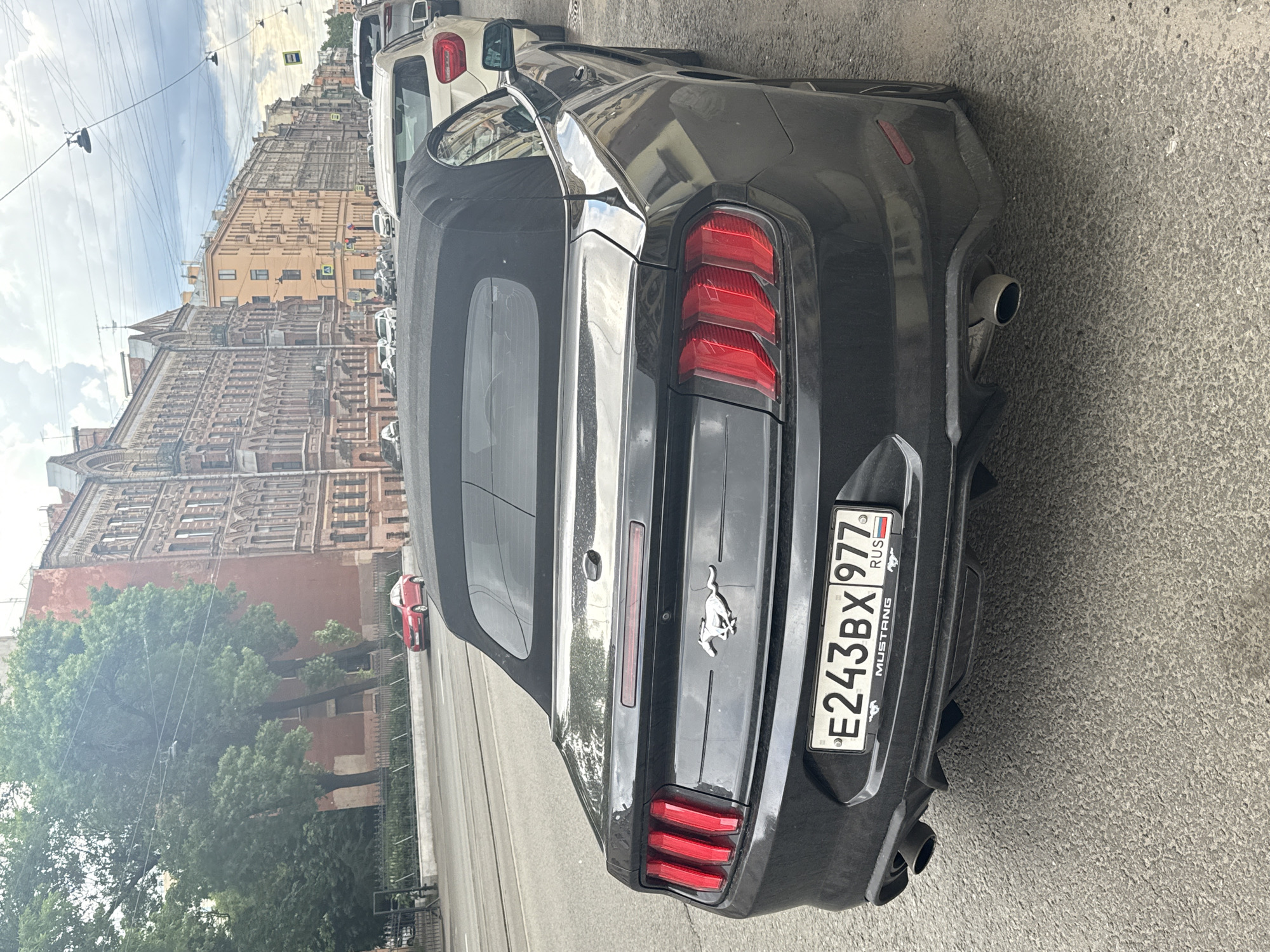 A black Ford Mustang convertible is parked on a city street in Saint Petersburg, Russia. The car is facing away from the camera, with its rear end visible. The license plate reads "E243BX 977" and has "RUS" written on it, indicating its Russian registration. The car has its black soft top up. Behind the Mustang, a white SUV is parked, and further down the street, several other cars and buildings are visible. The buildings appear to be multi-story residential or commercial structures with architectural details typical of Saint Petersburg. The street is paved with asphalt, and there are power lines and street lights overhead. The sky is overcast, suggesting a cloudy day. The scene appears to be a typical urban streetscape in a Russian city.