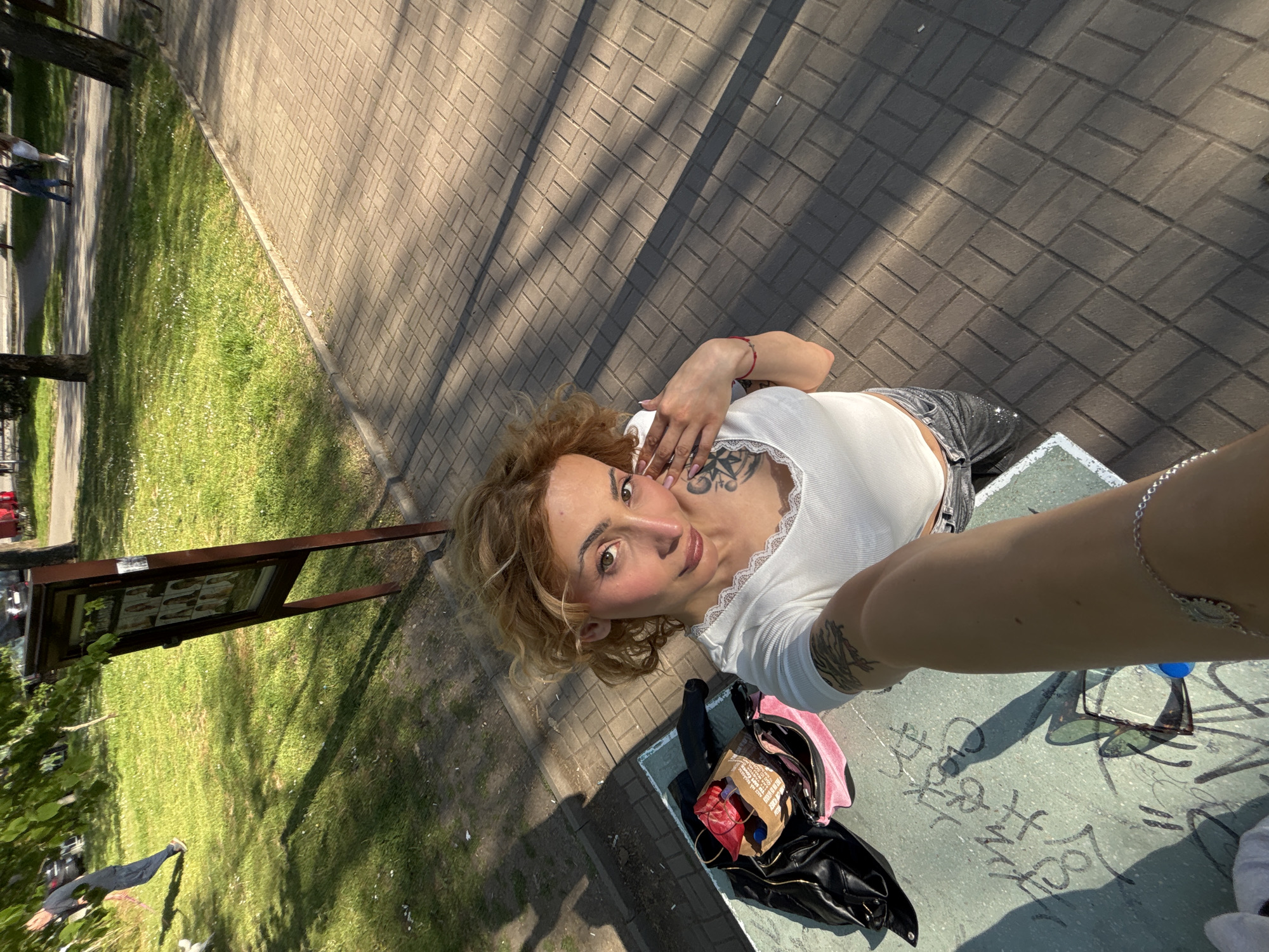 A medium shot from a high angle shows a fair-skinned person holding the camera at arm's length. The person has light brown, curly hair and is wearing a white, low-cut, ribbed top. They have multiple tattoos on their chest and arms. The setting appears to be a park or outdoor area within the city of Sofia, Bulgaria. The person is situated next to a low, concrete table with graffiti. Scattered on the table are personal items: a black jacket, a pink bag, a box, sunglasses, and a water bottle. The background includes a grassy area with trees, a brick-paved walkway, and an information board. The overall lighting suggests a sunny day.
