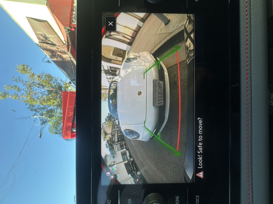 The image shows a car's backup camera display. A white Porsche is directly behind the vehicle, indicated by green lines on either side and a red line marking the closest distance. The display reads "Look! Safe to move?" along with a warning triangle. The display also shows options for PHONE, VOICE, RADIO, and MEDIA. The camera is showing the license plate of the Porsche.