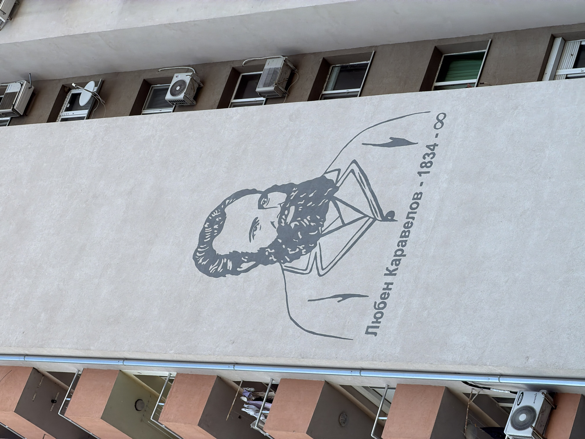 The image shows a section of a building in what appears to be the city of Blagoevgrad, Bulgaria. A mural of Lyuben Karavelov, a Bulgarian writer and revolutionary, is painted on the building's exterior wall, accompanied by the text "Любен Каравелов - 1834 - ∞". The building's architecture includes several windows with air conditioning units installed. The photograph is taken from a slightly elevated angle, revealing the building's facade and some of its lower features.