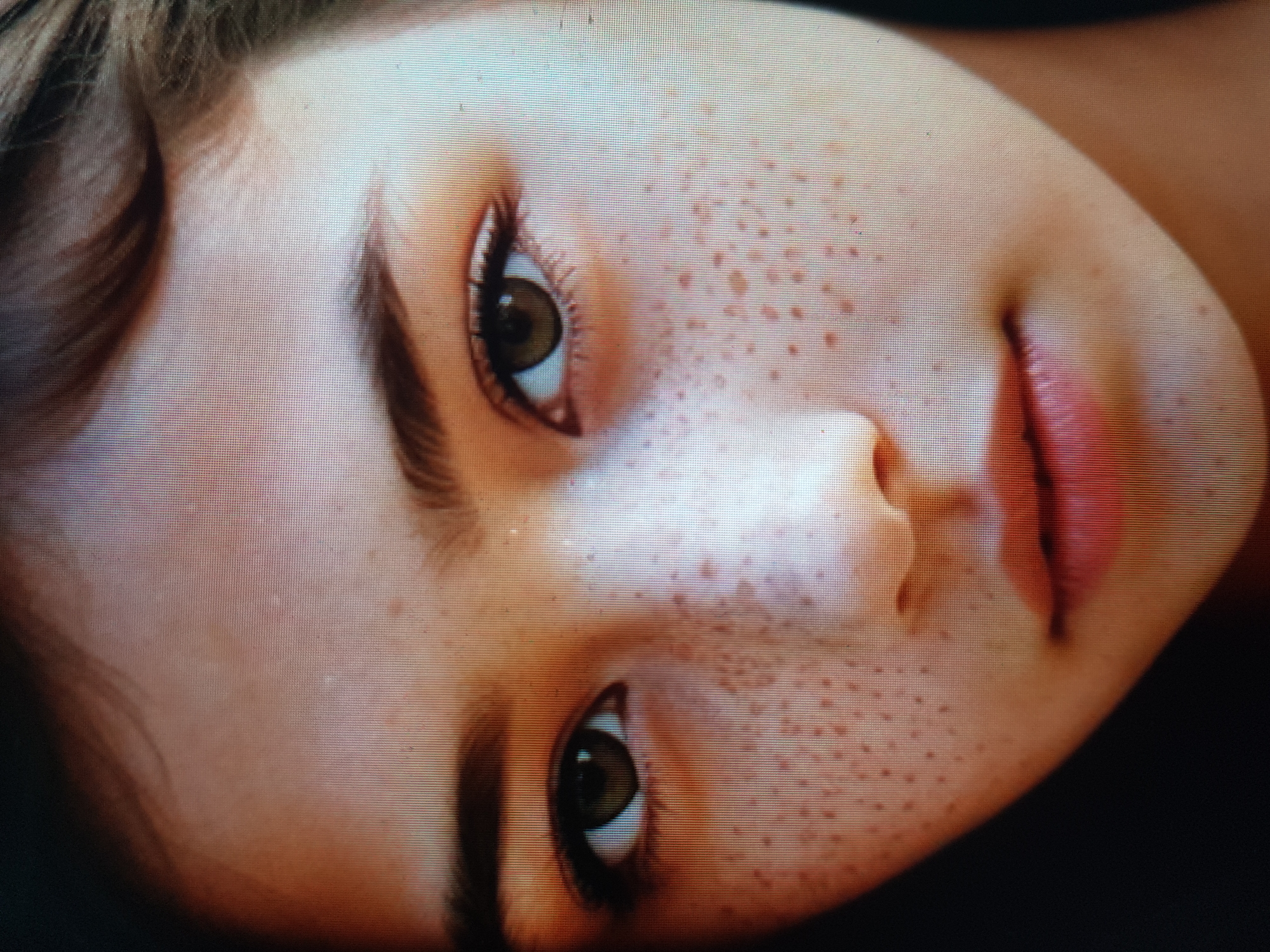 This is a close-up portrait of a young person's face. The person has light skin with numerous freckles scattered across their cheeks and nose. Their eyes are open and directed forward, with visible eyelashes and eyebrows. The iris color appears to be a light brown or hazel. The lips are slightly parted and have a pinkish hue. The hair on the forehead is dark brown and appears to be styled loosely. The background is dark and out of focus. There are no discernible elements that would indicate the location is Zherebkove, Ukraine.