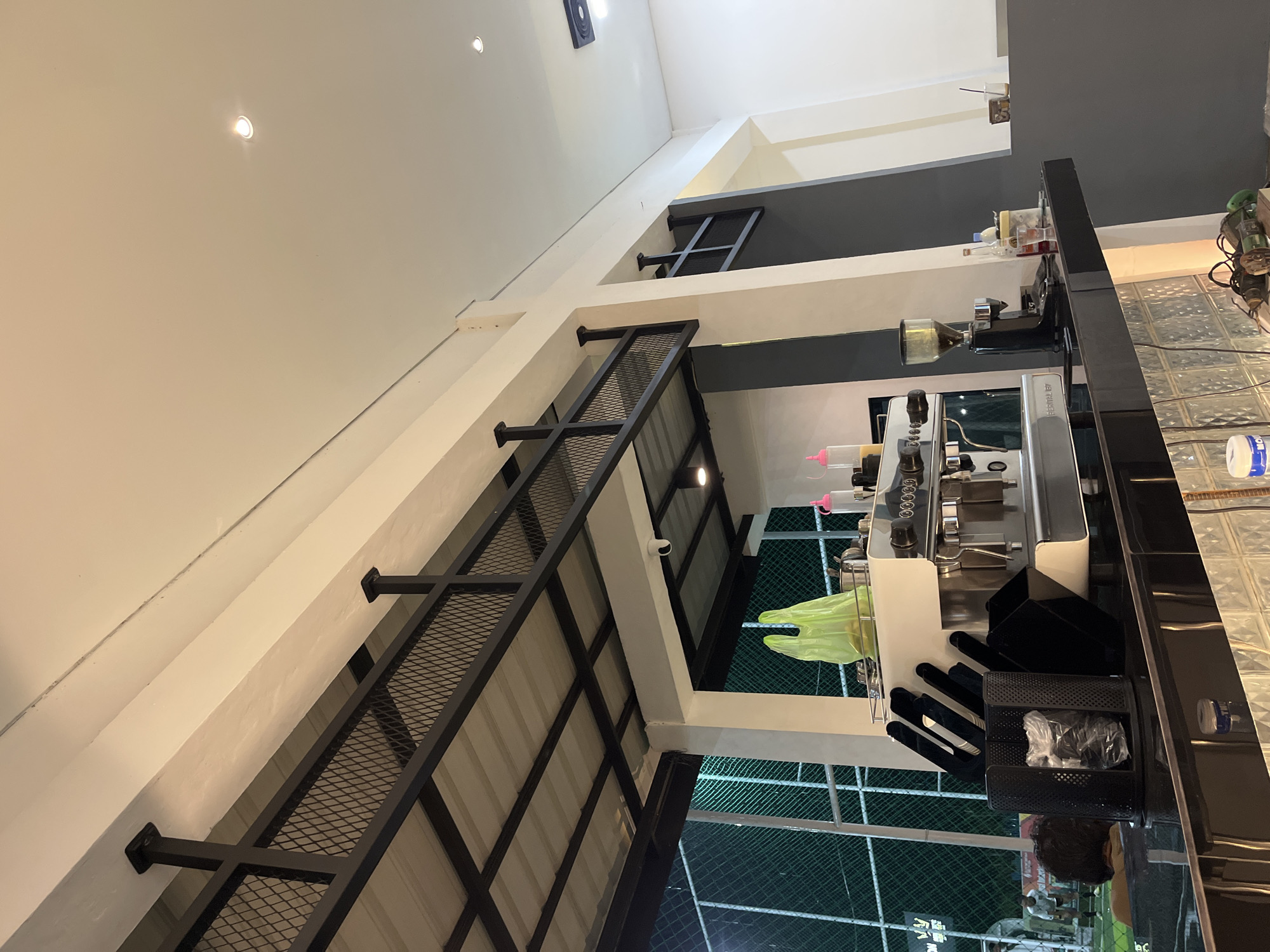 A slightly tilted, interior view shows a contemporary commercial space. The interior design includes a black metal grid structure extending from the ceiling. A service counter with various items, including an espresso machine and accessories, is visible. The background features a green, net-like pattern, suggesting an outdoor view or a decorative element. The overall location appears to be a commercial establishment in a modern setting. The location in the city of Lhokseumawe, Indonesia is not discernible from the image.