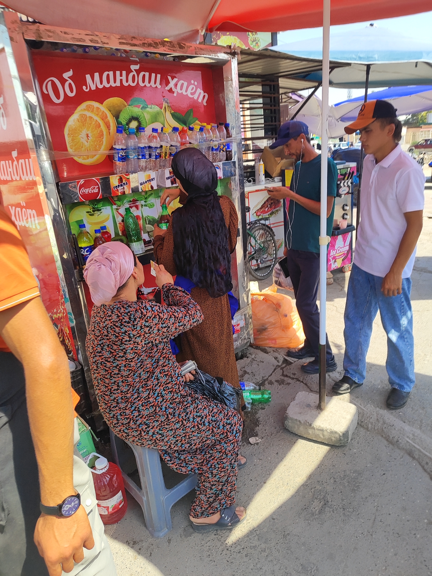 A street vendor is serving drinks from a cooler at a stall. The cooler is red and displays images of fruits, including oranges, kiwi, and apples. Several bottles of drinks are visible in the cooler, along with a large bottle of red juice on the ground. A woman wearing a pink headscarf and a floral dress is seated on a stool, holding a small item. Another woman in a dark headscarf and brown dress is standing behind the cooler, appearing to interact with the vendor. Two young men are standing near the cooler, one wearing a green shirt and dark pants, the other a white polo shirt and blue jeans. The background shows a street scene with umbrellas and other stalls. The sign above the cooler is in Cyrillic script, which is common in Tursunzoda, Tajikistan.