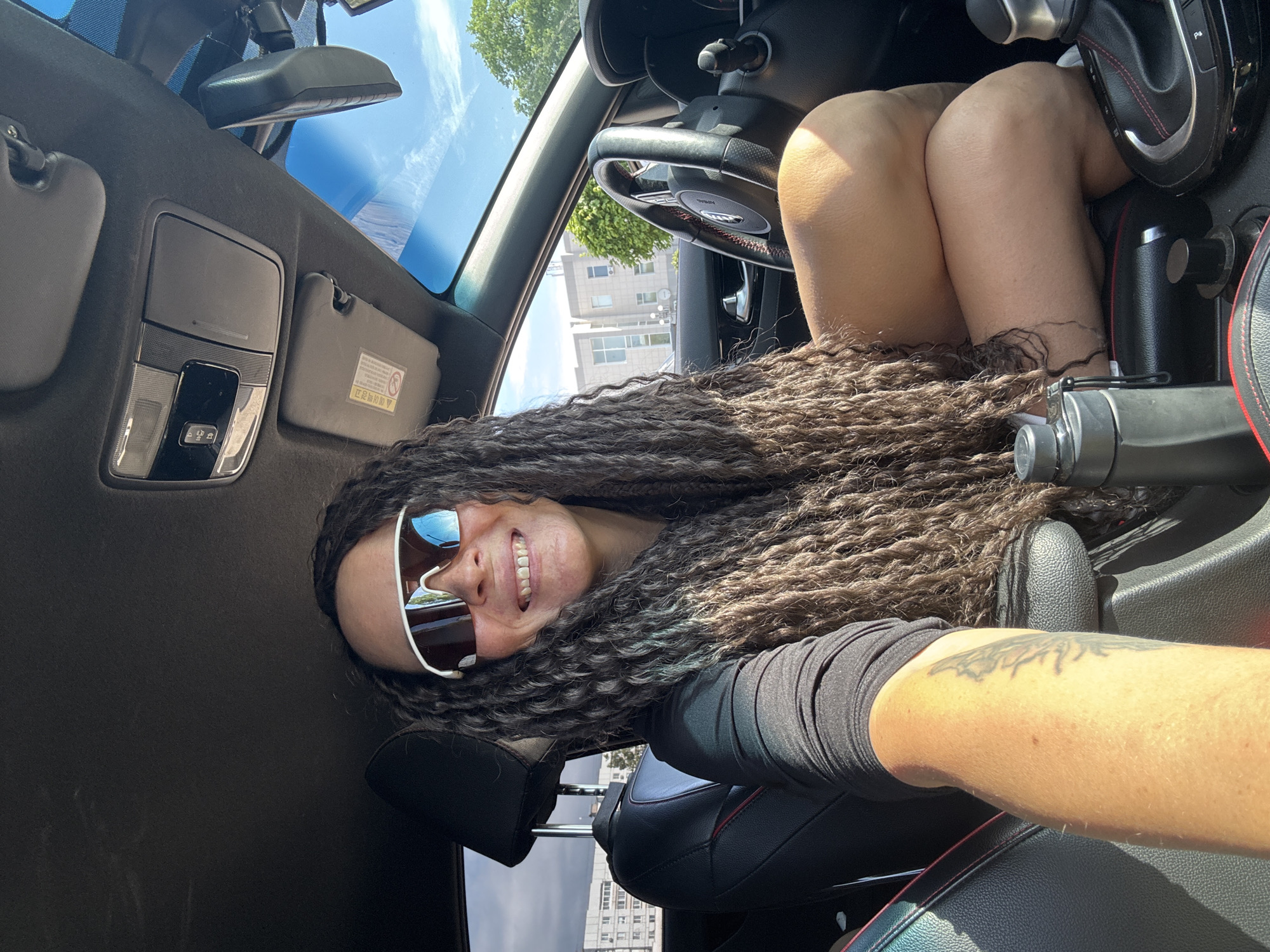 A woman is seated in the driver's seat of a car. The image shows her face, wearing sunglasses, and her hair is styled in long, twisted braids. Her knees are up near the steering wheel and dashboard. A portion of her right arm is visible and has a tattoo. The interior of the car is visible, including the steering wheel, dashboard, gear shift, and seats. Outside the car, a glimpse of a building is visible through the window. Given that the user indicated Chaikovsky, Russia, the building could be assumed to be located there.