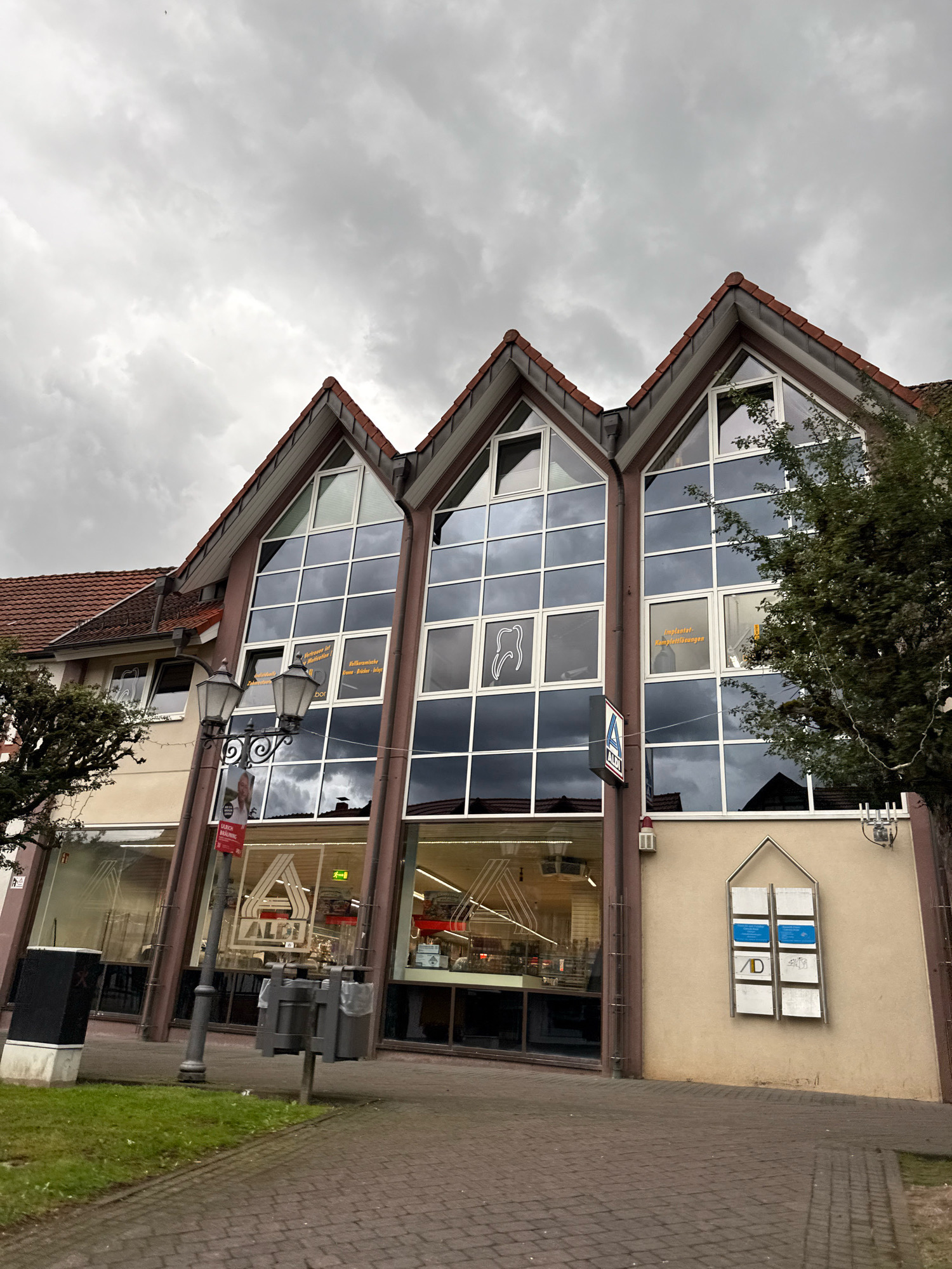 This is a wide shot, eye-level, outdoor, full, daylight, non-professional photograph of the Aldi supermarket in Bad Sachsa, Germany, on a cloudy day. The building has a unique architectural style with a series of gabled roofs and large windows that reflect the cloudy sky. A sign for Aldi is visible on the front of the building. To the left of the building, there is a lamppost with two lanterns and a sign that appears to be for a dentist. There is also a trash can and a patch of grass in the foreground. The street in front of the building is paved with brick.