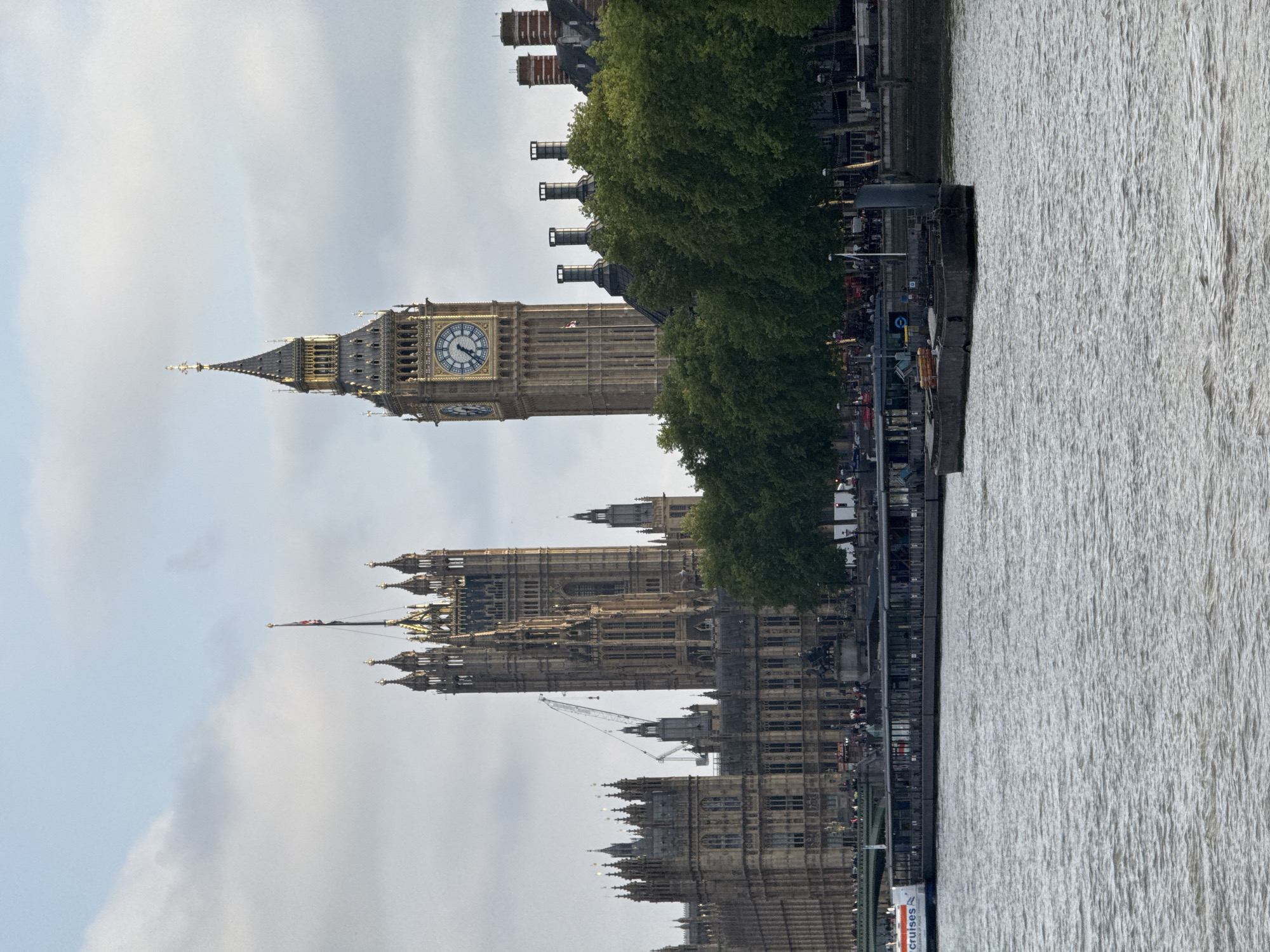This image shows the Palace of Westminster and Elizabeth Tower, commonly called Big Ben, in London, United Kingdom. The buildings are adjacent to the River Thames, which occupies the right side of the frame. People can be seen near the buildings. A flag flies atop the Palace of Westminster. The sky is overcast with clouds.