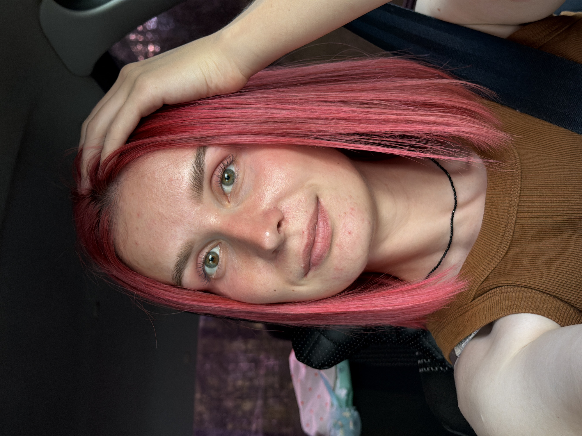 The image shows a close-up of a young woman with bright pink, shoulder-length hair. She is in the driver's seat of a car, as indicated by the seatbelt and dashboard visible. Her left hand is resting on top of her head, and she is looking directly at the camera with a slight smile. She is wearing a brown ribbed tank top and a thin black beaded necklace. The background is dark and out of focus, with some blurred lights suggesting foliage or other car lights outside the vehicle. There are no visible landmarks or indicators to determine a specific location within the city of Noviy, Russia.
